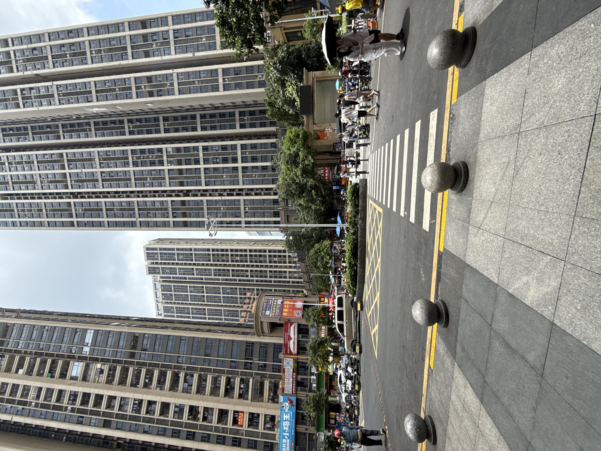 This is a street scene in Haizhu District, China. Tall, modern apartment buildings line the background. In the foreground, a crosswalk is visible on a paved street. Several people are crossing the street, some carrying umbrellas. A few motorcycles and a van are parked on the side of the road. The sky is overcast.