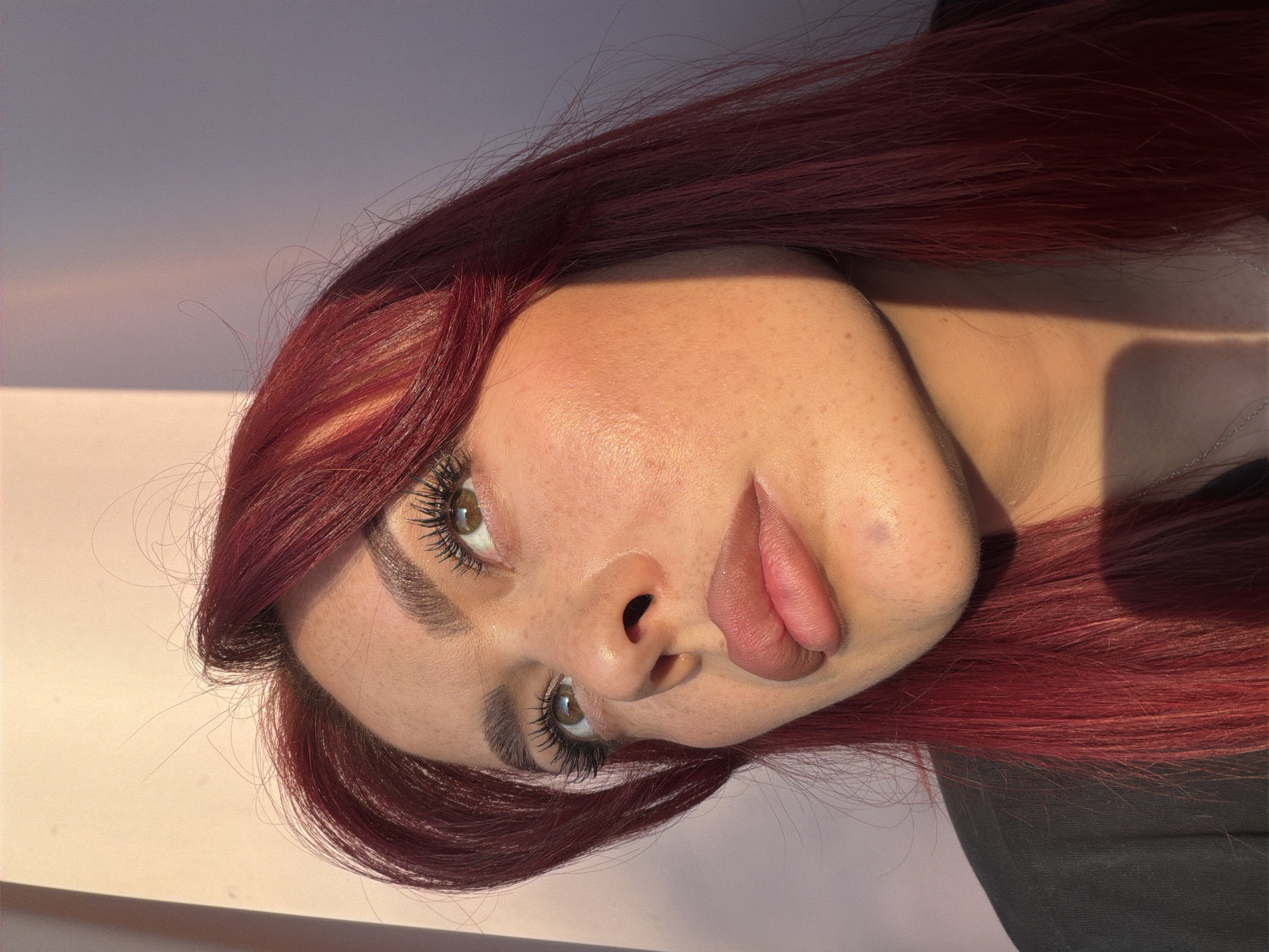 A close-up, low-angle portrait shows a woman with dark red hair and fair skin with freckles looking upwards. She is wearing a black top and has prominent eyelashes. The lighting suggests it is either sunrise or sunset, casting a warm glow on her face and creating a shadow line across her right cheek and neck. The background is a wall with a soft pink to purple gradient. The image does not contain any recognizable landmarks or elements that would indicate a specific location within the city of Uzukovo, Russia.
