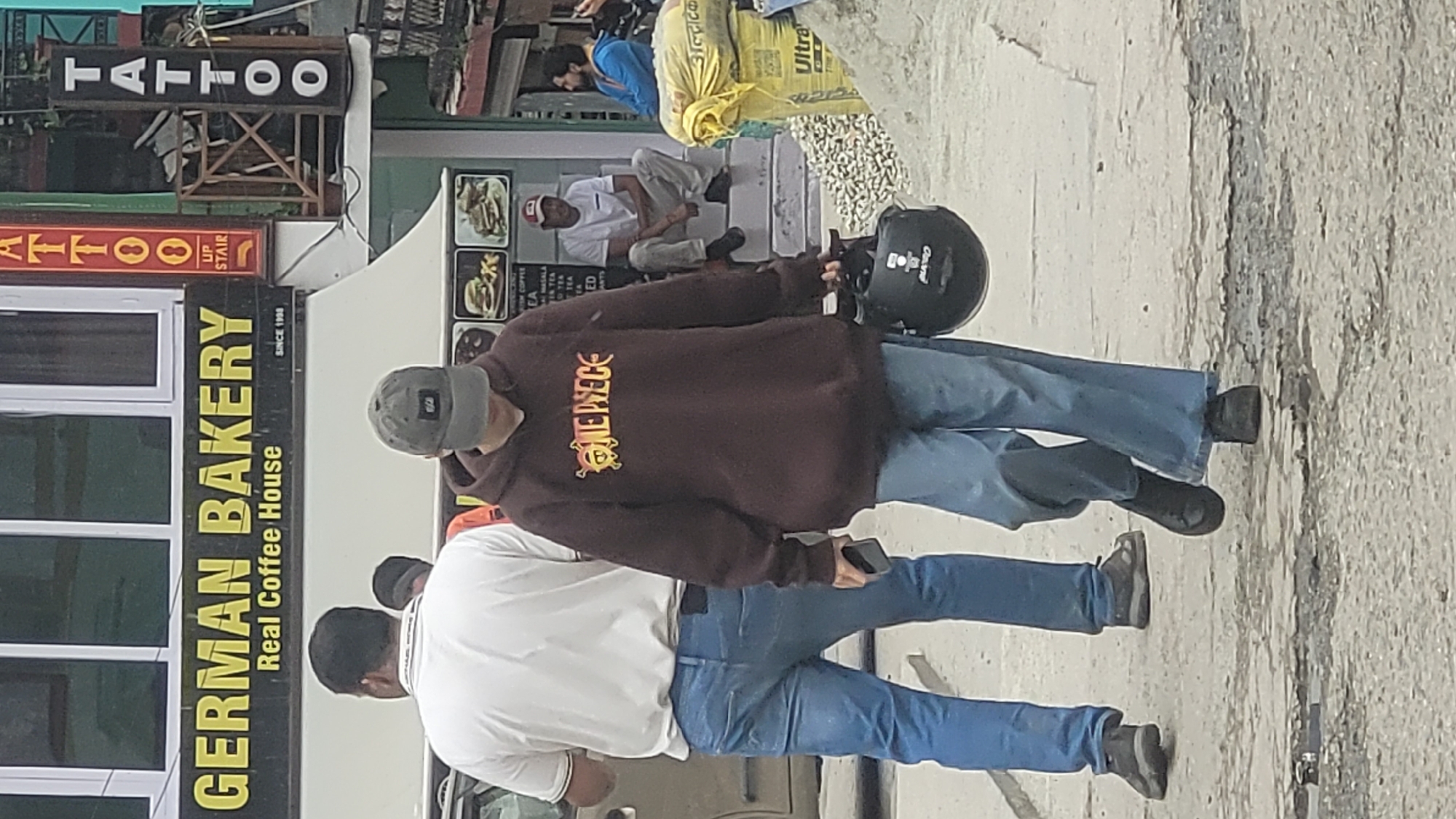 This image, taken in Vashisht, India, shows a street scene with several people and commercial signs. In the foreground, two individuals are partially visible, with one wearing a brown hoodie with "ONE PIECE" written on it and the other a white shirt. Both are wearing blue jeans. A black helmet rests on the ground near the person in the hoodie.
Behind them, a large sign reads "GERMAN BAKERY Real Coffee House SINCE 1998" in prominent yellow letters. Other signs visible include one for "TATTOO" and another for "ATTOO" with "UP STAIR" indicated. There are also images of food items, likely from a cafe or restaurant menu.
In the background, other people are present, including someone wearing a blue shirt and another sitting down. Bags of what appear to be cement or construction material are also visible, suggesting a somewhat busy or developing area. The overall impression is of a commercial street in a populated area. The lighting suggests it is daytime.