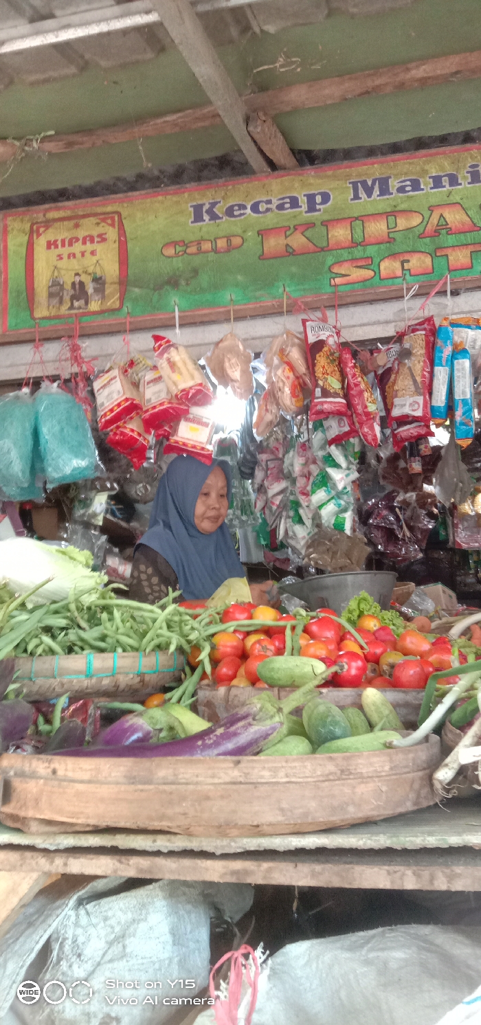 This image depicts a market stall in Maron, Indonesia. An elderly woman, wearing a blue hijab and a patterned dark shirt, is seated behind a counter laden with fresh produce. Her gaze is directed slightly to her right, away from the camera.
In the immediate foreground, a large, shallow, round wooden tray or basket holds a variety of fresh vegetables, including multiple purple eggplants, green cucumbers, and green beans. Below this tray, a rustic wooden shelf is visible, and further down, large light-colored sacks are present.
On the main sales counter behind the foreground tray, numerous piles of produce are displayed. Prominent items include bright red and orange tomatoes, green beans, and lettuce. Behind the woman, numerous packaged items, including snacks and dry goods, are hung from lines and displayed on shelves, obscuring the background. Some visible packaging suggests various food products, including items labeled "ROMSYO".
Above the entire scene, a large banner is displayed, partially visible, featuring text in Indonesian. The banner reads "Kecap Manis Cap Kipas Sate" (Sweet Soy Sauce, Kipas Sate Brand) and features a smaller graphic panel on the left with the text "KIPAS SATE" and an illustration of two figures with shoulder poles. The stall's ceiling consists of rough wooden beams and a green painted surface. A watermark for "Shot on Y15 Vivo AI camera" is present in the lower-left corner.