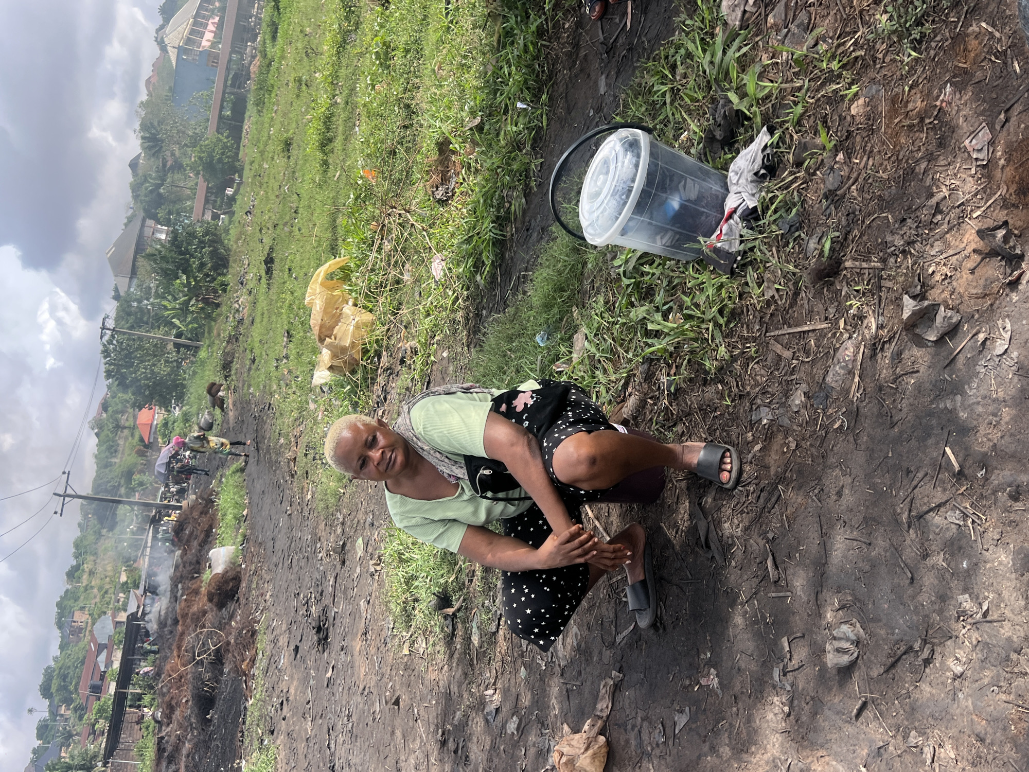 In an outdoor environment in Obosi, Nigeria, a woman is crouched on the ground, appearing to examine her feet. She wears a light green top, a black skirt with white stars, and gray sandals. A transparent plastic bucket is located to her right. The ground appears muddy and soiled, with remnants of burning debris. In the background, there are houses, a power pole, and other people. There is smoke in the distance, suggesting a recent fire. The sky is overcast.