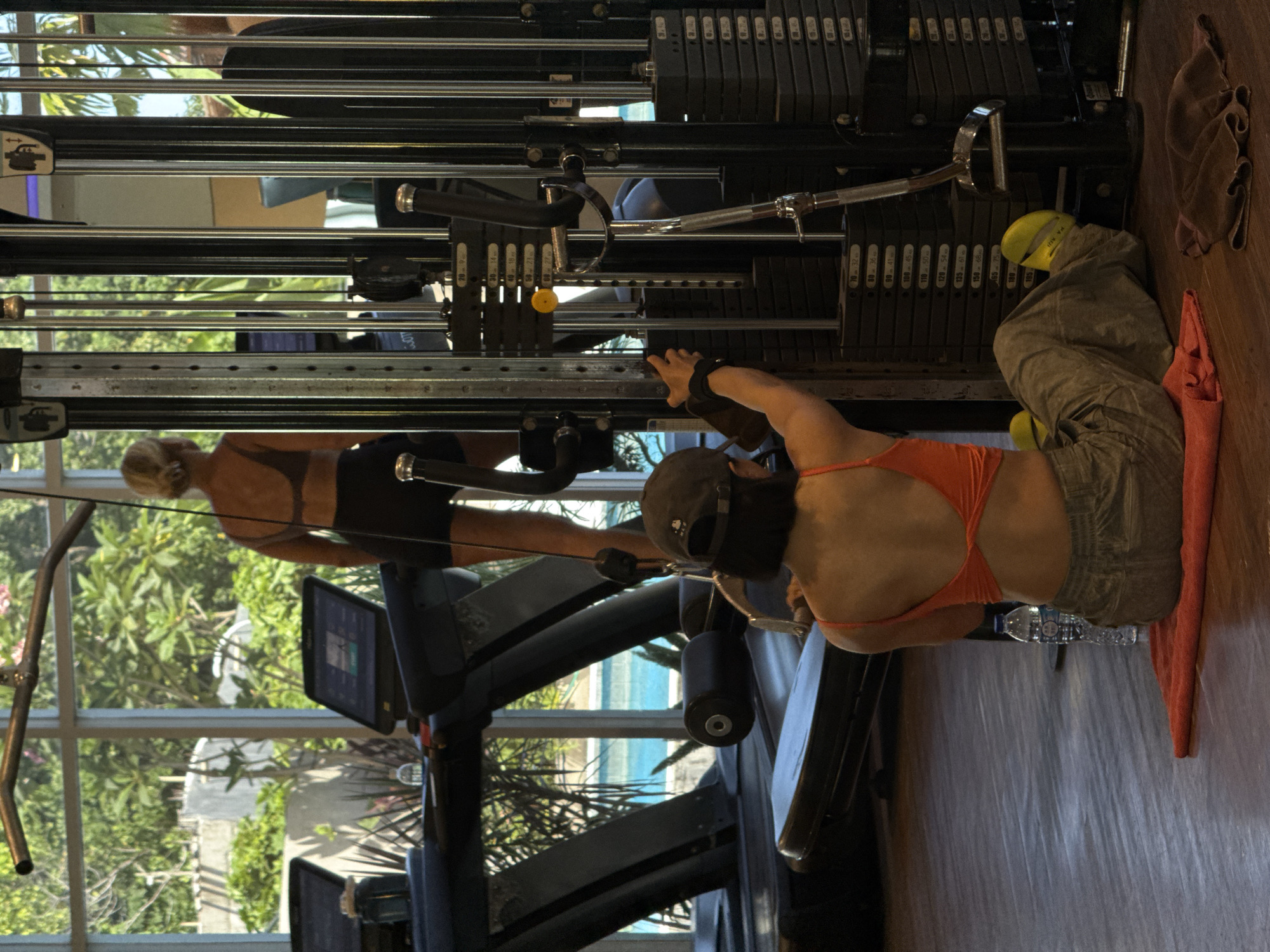 The image captures a fitness scene within a gym, likely located in North Kuta, Indonesia, based on the apparent tropical foliage and building exteriors visible through the window.
On the right side of the frame, a person wearing an orange sports bra, grey pants, and a dark baseball cap is positioned in front of a weight machine. They appear to be engaged in a workout, pulling the machine's cables. A water bottle rests on the floor beside them, near an orange towel.
In the background, visible through the gym equipment's framework, another individual in athletic attire, consisting of a dark sports bra and shorts, is exercising on a treadmill.
The gym is well-lit, with large windows offering a view of outdoor greenery, suggesting the location of North Kuta in Indonesia.