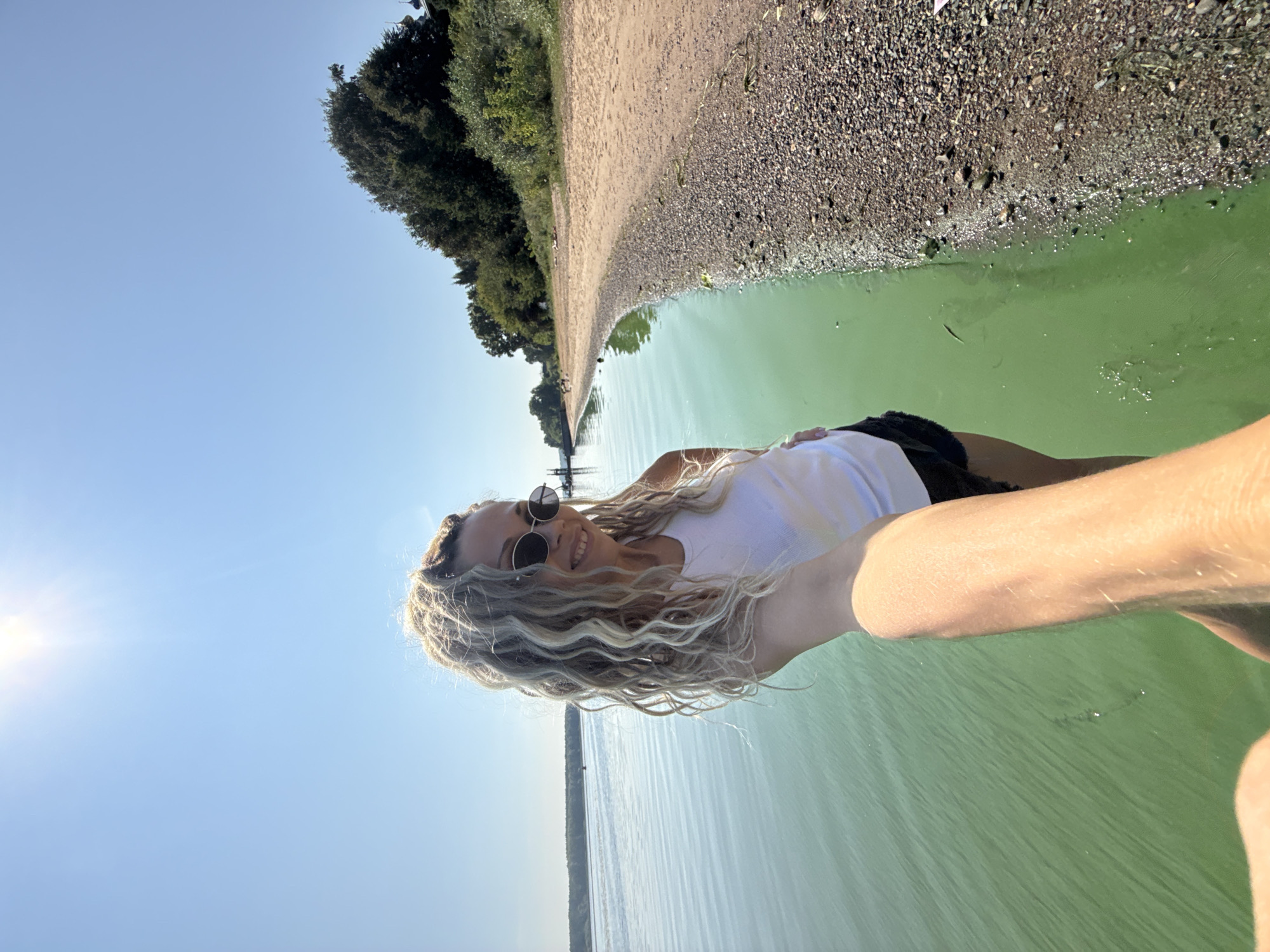 A woman with blonde, curly hair and sunglasses is taking a selfie on a sunny day at the shore of a body of water, likely a lake or river, in Rusanovo, Russia. She is wearing a white tank top and black shorts. The water is a murky green color and the shoreline is composed of sand and small pebbles. In the background, there are trees and vegetation along the bank, and a clear blue sky overhead. The sun is shining brightly, casting a glare on the water and sky.