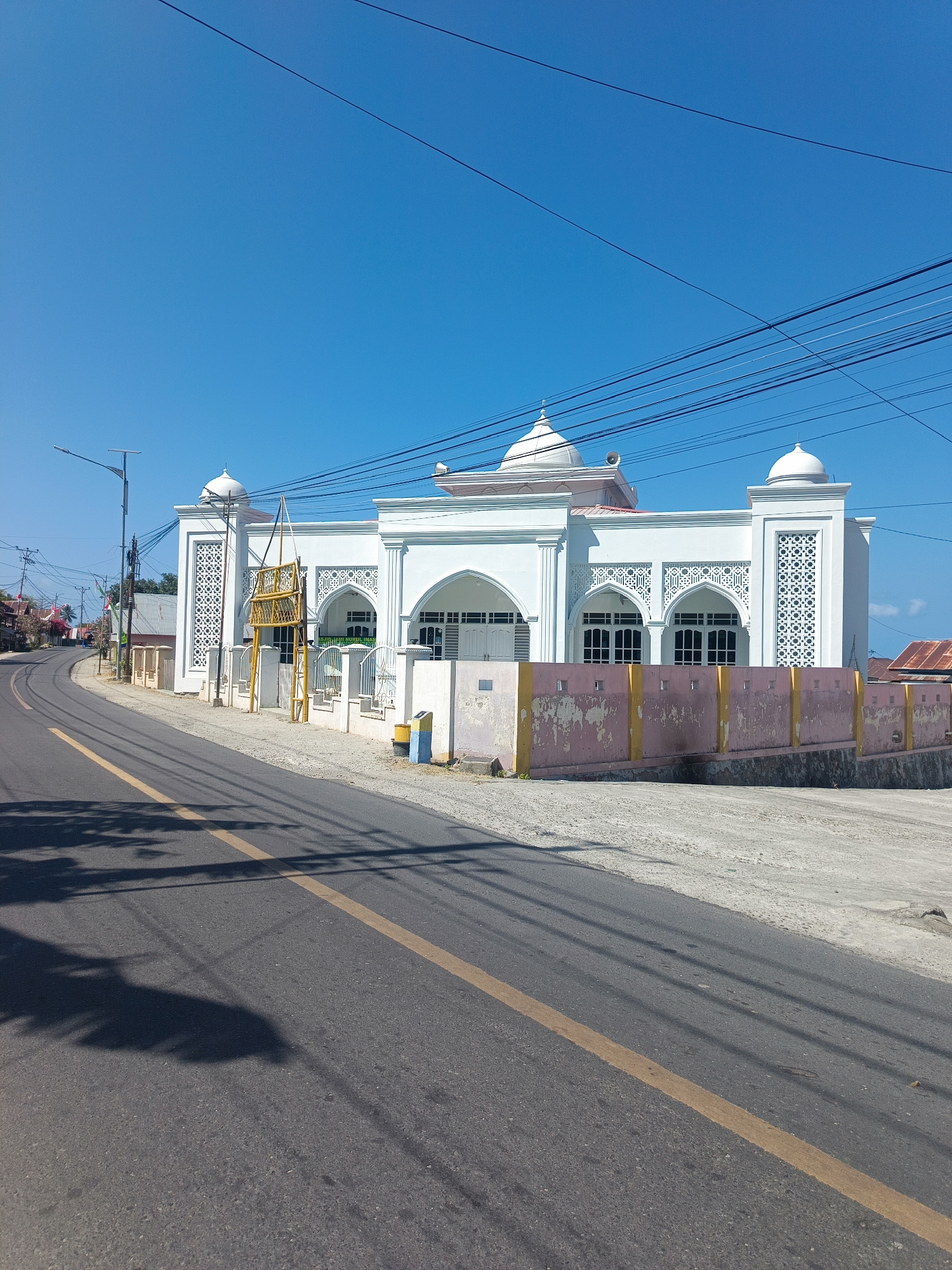 The image shows a white mosque with a dome, located on the right side of a paved road. The mosque features intricate white latticework on its facade and is surrounded by a low, pink and yellow painted wall. A street lamp is visible on the left side of the road, and utility wires crisscross the sky above. The road is asphalt with a yellow line down the center, and shadows of trees are cast on its surface. The sky is clear and blue. The location is identified as Huangobotu, Indonesia.