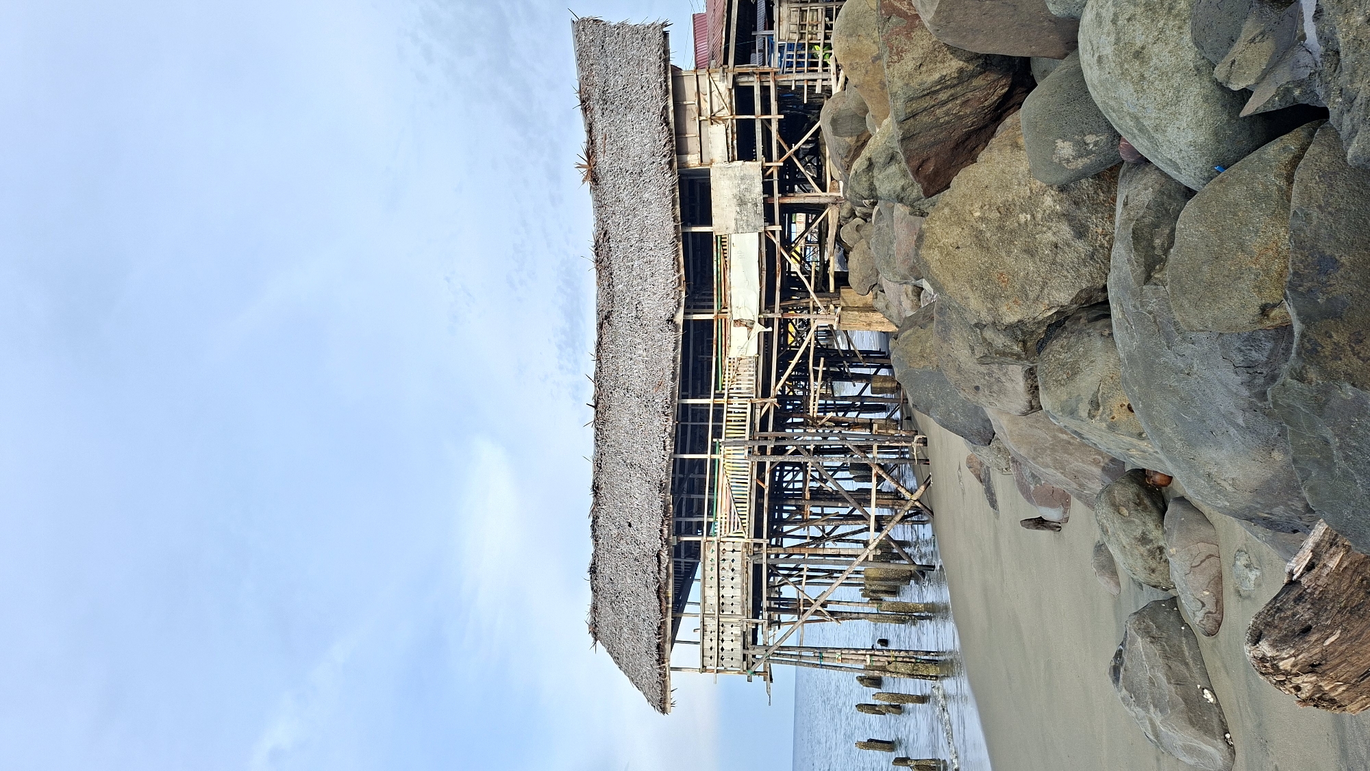 A wooden stilt house with a thatched roof sits on the beach in Lhokseumawe, Indonesia. The house, which appears to be a restaurant or a similar establishment, is supported by numerous wooden stilts that extend into the ocean. The structure shows signs of age and weathering, with some parts appearing unfinished or in disrepair. In the foreground, a pile of large rocks and driftwood lines the sandy beach. The ocean is calm, with gentle waves lapping the shore. The sky is overcast with a light blue hue. No people are visible in the image. The overall scene depicts a rustic and weathered beachfront structure in a coastal setting.