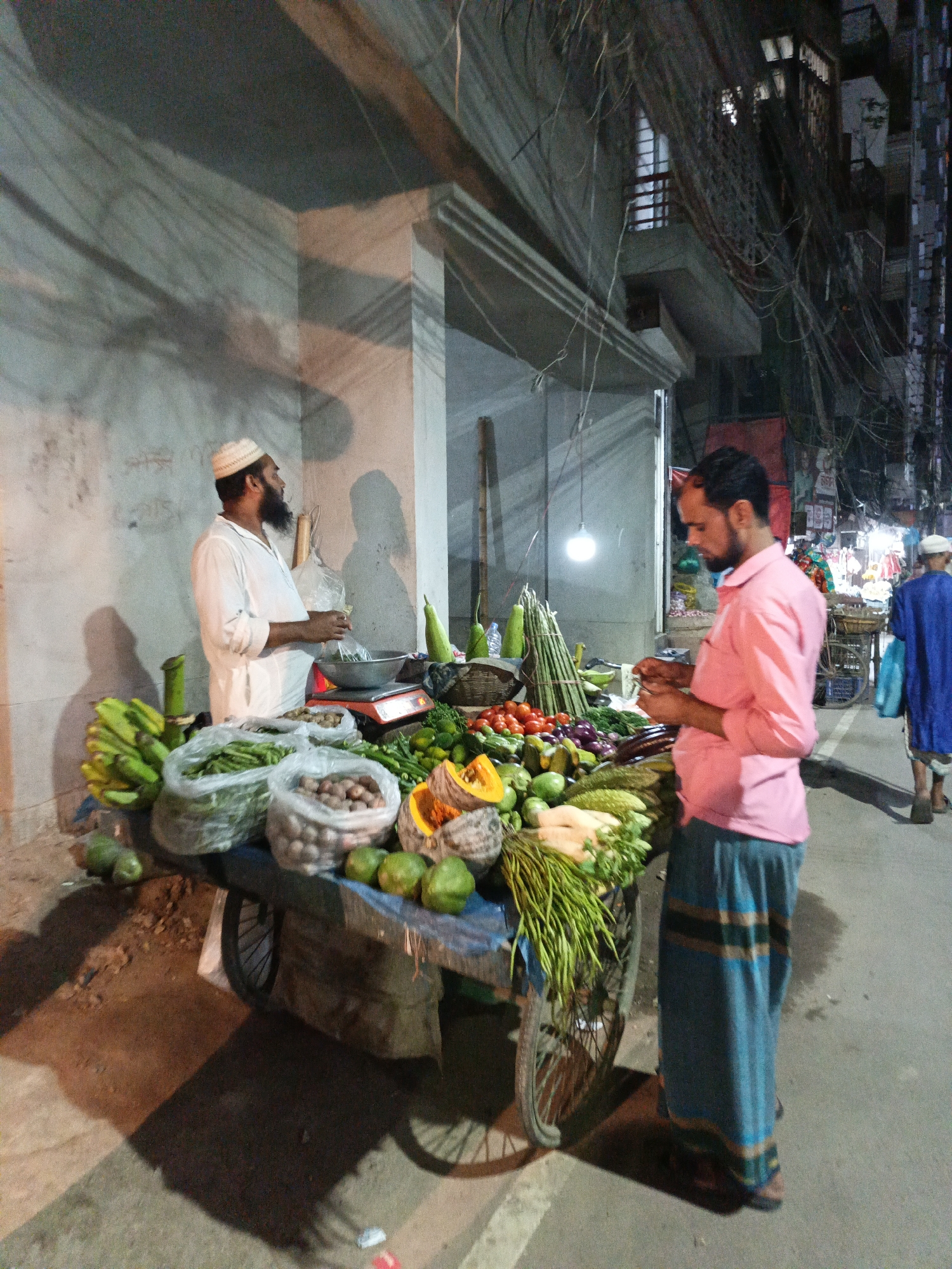 The image captures a vibrant nighttime street scene in Dhaka, Bangladesh, centered around a mobile vegetable stall. Two men are engaged in what appears to be a market transaction.
On the left, a vendor, dressed in a white long-sleeved tunic and a white skullcap, stands behind his cart. He has a beard and is looking towards the customer, with a weighing scale (red base, silver pan) and a plastic bag visible on his stall. His shadow is cast prominently on the light-colored wall behind him.
Facing him is a customer on the right, wearing a pink collared shirt and a blue patterned lungi. He also has a beard and is looking down, seemingly inspecting the produce or his hands.
The wheeled cart is heavily laden with a diverse selection of fresh fruits and vegetables. Visible items include bunches of green bananas, bags of potatoes, large green gourds, ripe red tomatoes, eggplants, limes, bundles of long green beans, and cut pumpkins or squash. A single bare light bulb hangs above the cart, illuminating the produce and creating distinct shadows on the ground and surrounding walls, indicative of the nighttime setting.
In the background, another man in a blue garment is partially visible walking away on the right. The urban environment features concrete buildings with windows and balconies. Overhead, a dense and complex network of tangled electrical and communication wires crisscrosses the sky, a characteristic feature of cities like Dhaka. The ground is a mix of asphalt and dirt. The scene portrays a common street market activity, likely after dark. No discernible text is visible.