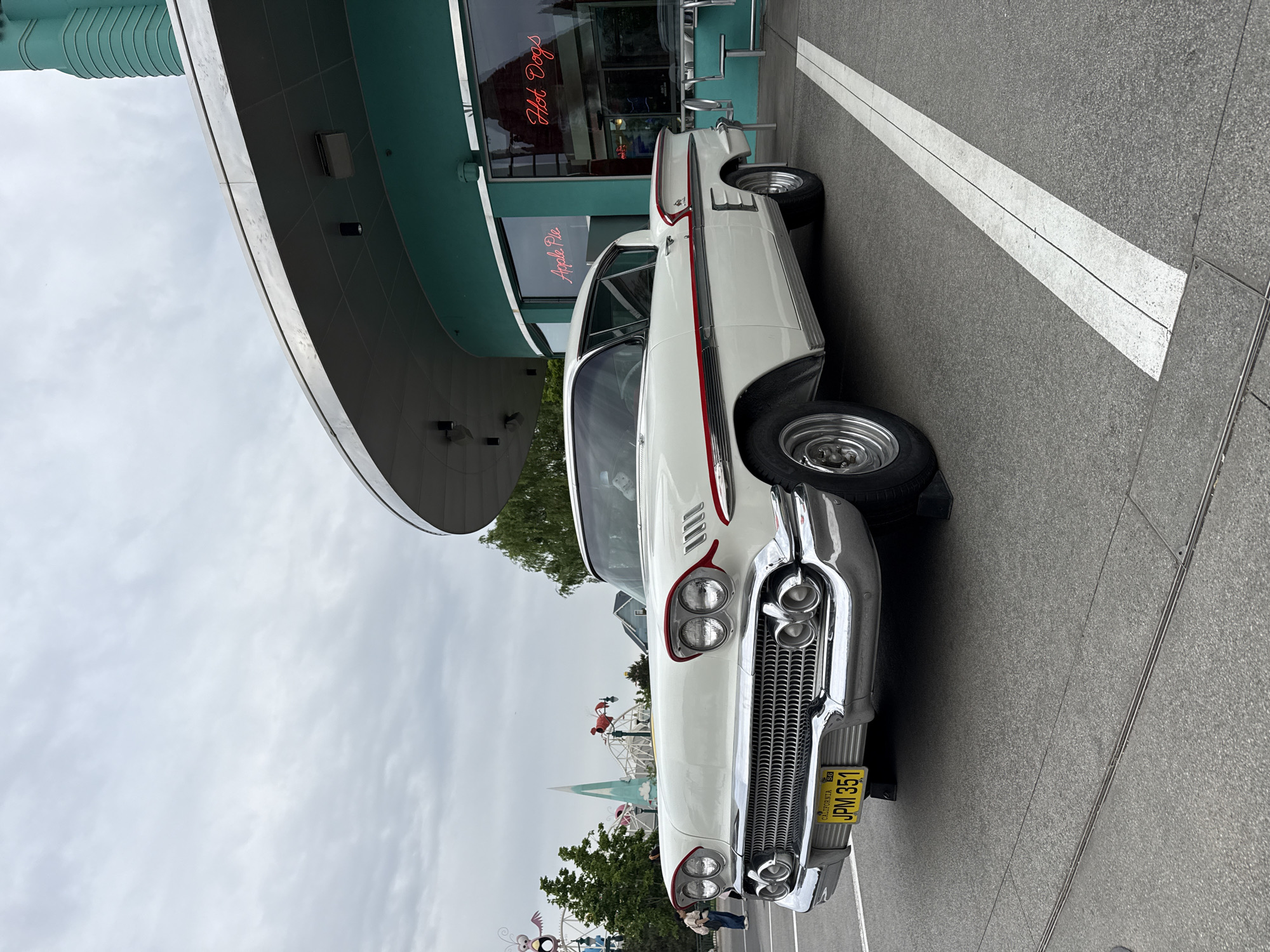 The image shows a vintage, white 1958 Chevrolet Impala parked in front of a diner with "Hot Dogs" and "Apple Pie" neon signs. The car has a red trim and a yellow California license plate reading "JPM 351". In the background, there's a park with a Ferris wheel and other amusement park rides. The location is outside of Tongzhou District, China. The sky is overcast. A white line is painted on the ground next to the vehicle.