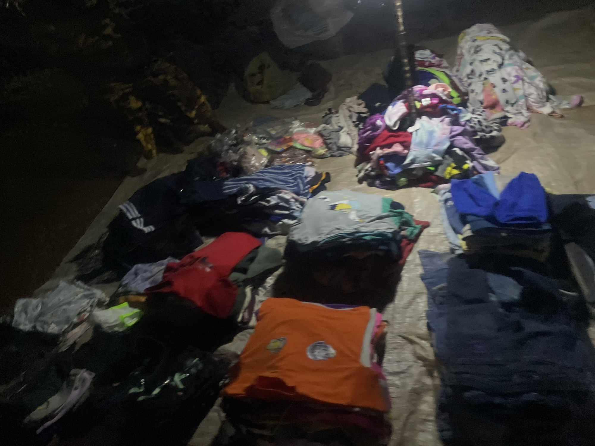 The image shows a pile of clothing laid out on the ground, likely as part of a market stall or a distribution event. The clothing appears to be a mix of casual wear, including t-shirts, pants, and possibly some pajamas or blankets, in various colors and patterns. Some items are folded, while others are more loosely piled. The scene is dimly lit, suggesting it might be evening or nighttime, or an indoor space with poor lighting. The ground cover appears to be a tarp or similar material. The overall impression is one of abundance and a variety of items for sale or giveaway. There are no people clearly visible in the frame, and no discernible text on the clothing. The location context provided indicates Sauka, Nigeria, which helps to understand the setting as an outdoor market or community gathering in that region. The image is somewhat blurry due to low light conditions and potentially motion.