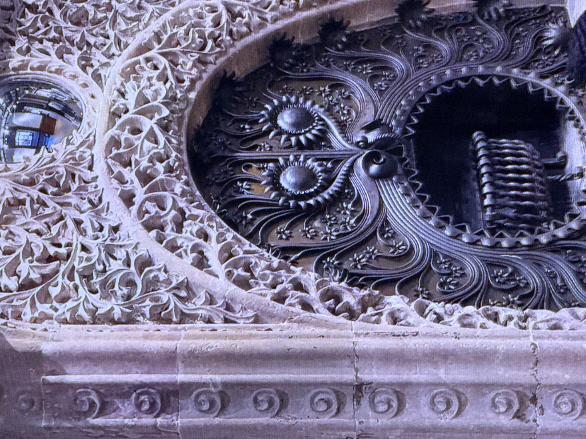 This is a close-up, medium shot of an ornate, dark metal fireplace surround, likely made of wrought iron or a similar material, featuring intricate scrolling designs, floral motifs, and two prominent sun-like circular embellishments that resemble eyes. The fireplace opening itself is dark and appears to contain a metal grate. To the left of the fireplace, there is a stone column with carved spiral designs. The background shows a highly decorative, light-colored carved stone archway above the fireplace, with a circular reflection visible within it, possibly of a room. The lighting appears to be interior. The image does not depict the city of Dubai or the United Arab Emirates.