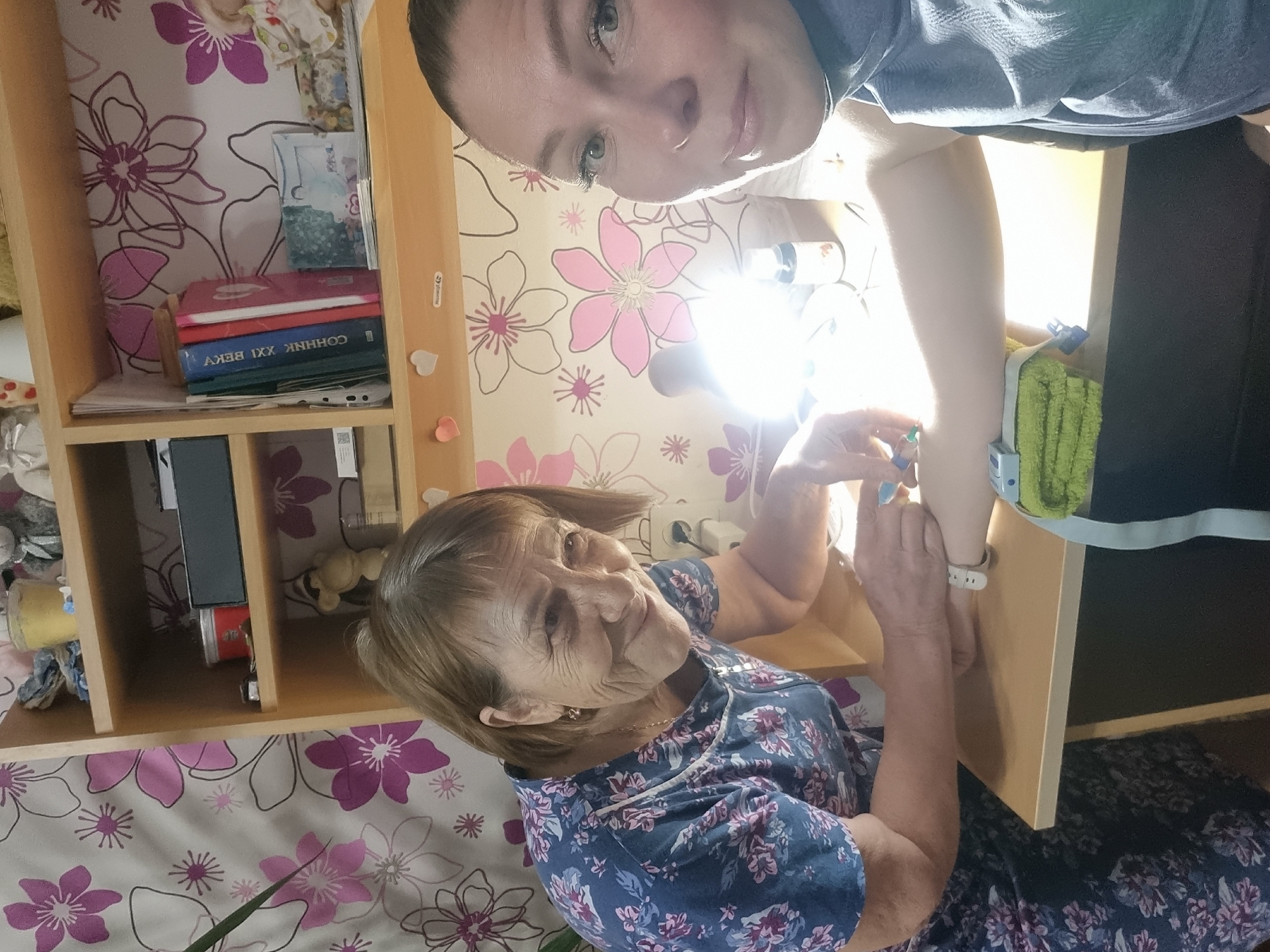 This image, taken in Chaikovsky, Russia, captures a tender interaction between two women in what appears to be a home setting. The photograph is oriented sideways, with subjects appearing rotated 90 degrees clockwise.
In the upper part of the frame, a younger woman looks directly at the camera with light eyes and her hair pulled back. She is wearing a grey/blue t-shirt. Her right forearm extends downwards, where a white medical device, possibly a brace or cuff, is visible, partially covered by a green towel. She holds a small vial or dropper in her left hand, appearing to be administering something to the older woman's hand or arm. A white smartwatch is visible on her wrist.
In the lower part of the frame, an older woman with wrinkles on her face and short, light brown hair looks up at the younger woman with a gentle, appreciative smile. She wears a dark blue dress adorned with a floral pattern of pink and purple flowers. Her hands are gently placed around the younger woman's forearm, suggesting a moment of care or assistance.
The background features a wooden shelving unit containing several books, one of which has the Cyrillic text "СОЧИННИК XXI ВЕКУ" (Sochinnik XXI Veky), translating to "Compositions of the XXI Century." Small, decorative heart-shaped magnets are attached to the side of the shelf. The walls are covered with wallpaper showcasing a pattern of large pink and purple flowers with outlined details. A bright light source, likely a lamp or flash, creates a prominent glare in the center of the image, illuminating the scene. A wall outlet with a plug is also visible. The overall atmosphere conveys a moment of affectionate care.