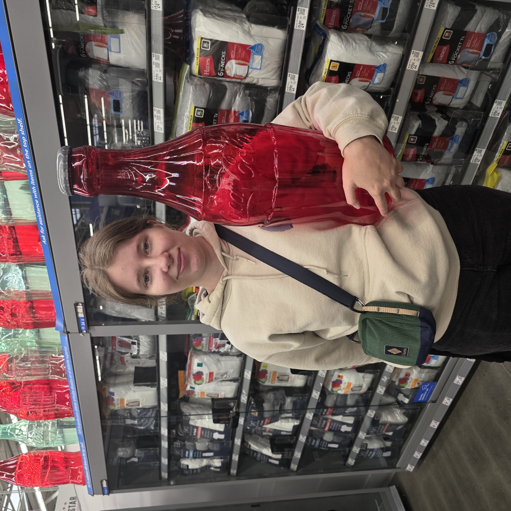 A young woman stands in an aisle of a retail store, holding a large, red, novelty Coca-Cola bottle. She is wearing a light tan hooded sweatshirt and dark jeans, with a green crossbody bag. The shelves behind her are stocked with packaged t-shirts, primarily Hanes brand. The store appears to be a large general merchandise retailer, potentially a Walmart or similar supercenter, given the type of products and store layout. The lighting is bright and artificial, typical of an indoor retail environment. The background also shows decorative, oversized Coca-Cola bottles in red and green. This store is located in Tigard, Oregon, United States of America.