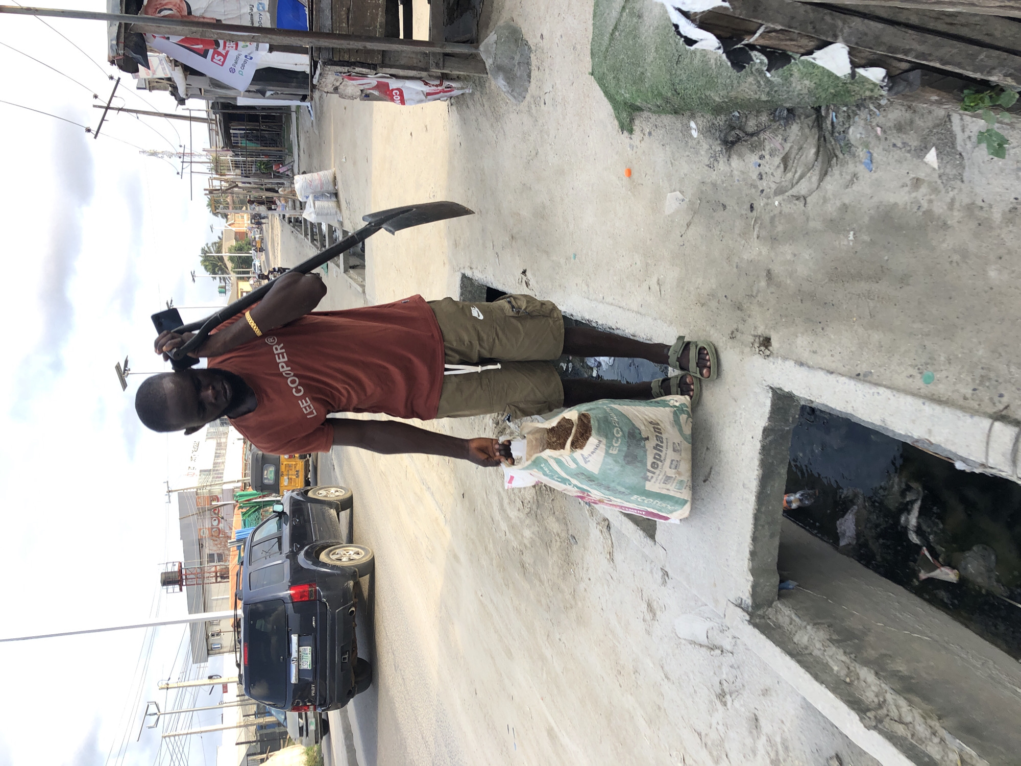 A man stands by a concrete drainage ditch on a street in Ado, Nigeria. He is dark-skinned, wearing a reddish-brown t-shirt with "LEE COOPER" printed on it, tan shorts, and sandals. He holds a white sack, printed with "Elephant Cement" and "ECO Plane", and a black shovel is positioned over his shoulder. The ditch is filled with dark water and various pieces of debris. To the left, a black Honda Pilot with license plate "LSD-774 HR" is parked on the side of the road. In the background, there are buildings, poles with electrical lines, and other cars on the street. A sign with "lodo d" and "aimPay" is also visible in the background. The sky is overcast.