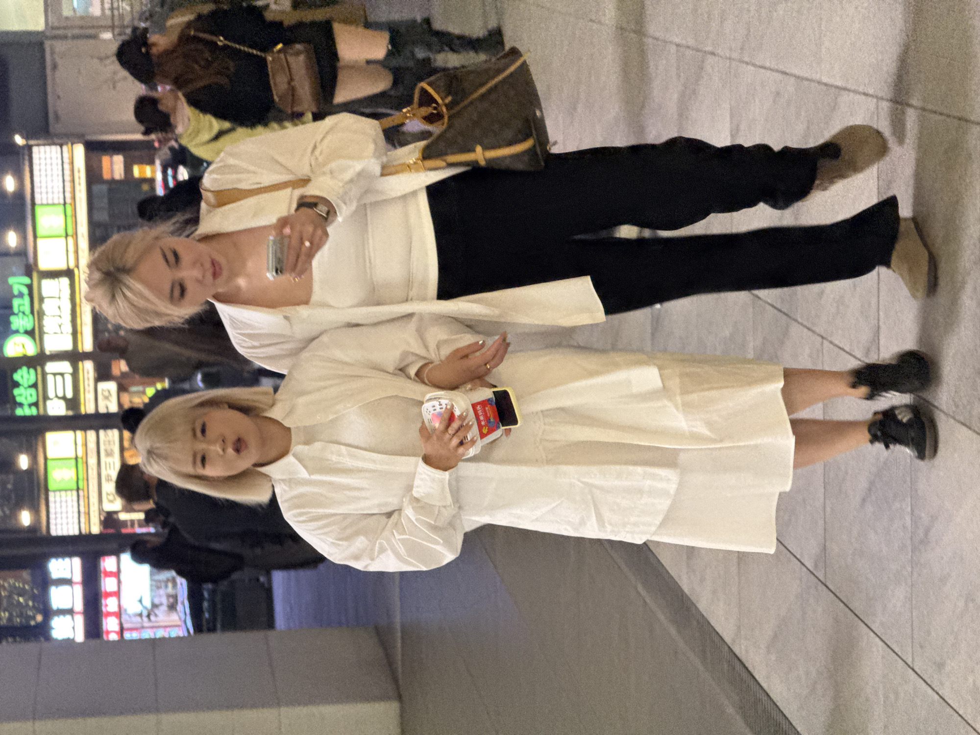 Two women with blonde hair walk side-by-side on a paved sidewalk at night in the city of Liangjiang, China. The woman on the left has a bob haircut, wears a white collared long-sleeve shirt over a white tiered midi dress, and black sneakers. She holds a clear plastic cup with red and white contents and a yellow-cased smartphone in her right hand. Her mouth is slightly open, suggesting speech. The woman on the right has her blonde hair pulled back, wears a white collared long-sleeve shirt over a white tank top, black pants, and tan flat shoes. She holds a silver-colored smartphone in both hands, looking at its screen. A dark wristwatch is visible on her left wrist, and she carries a brown monogrammed shoulder bag. Her left arm appears to be linked with the right arm of the woman on the left. In the blurred background, other pedestrians are visible, along with brightly lit commercial buildings displaying various signs in Chinese and Korean characters.