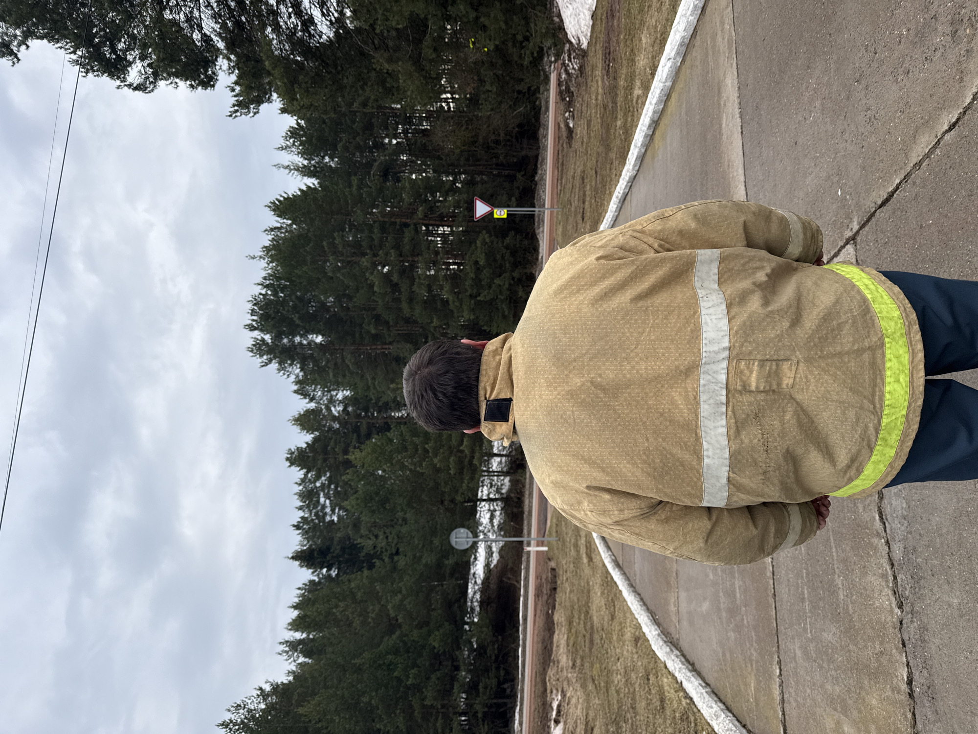 A person wearing a tan jacket with reflective stripes stands on a paved path, likely a sidewalk, in an outdoor environment. The person faces a forest area, with a road sign visible in the distance. The background consists of a cloudy sky and tall trees. The location is presumed to be in or near Rusalevka, Russia. The individual is wearing a tan jacket and dark pants and standing on a grey concrete road.
