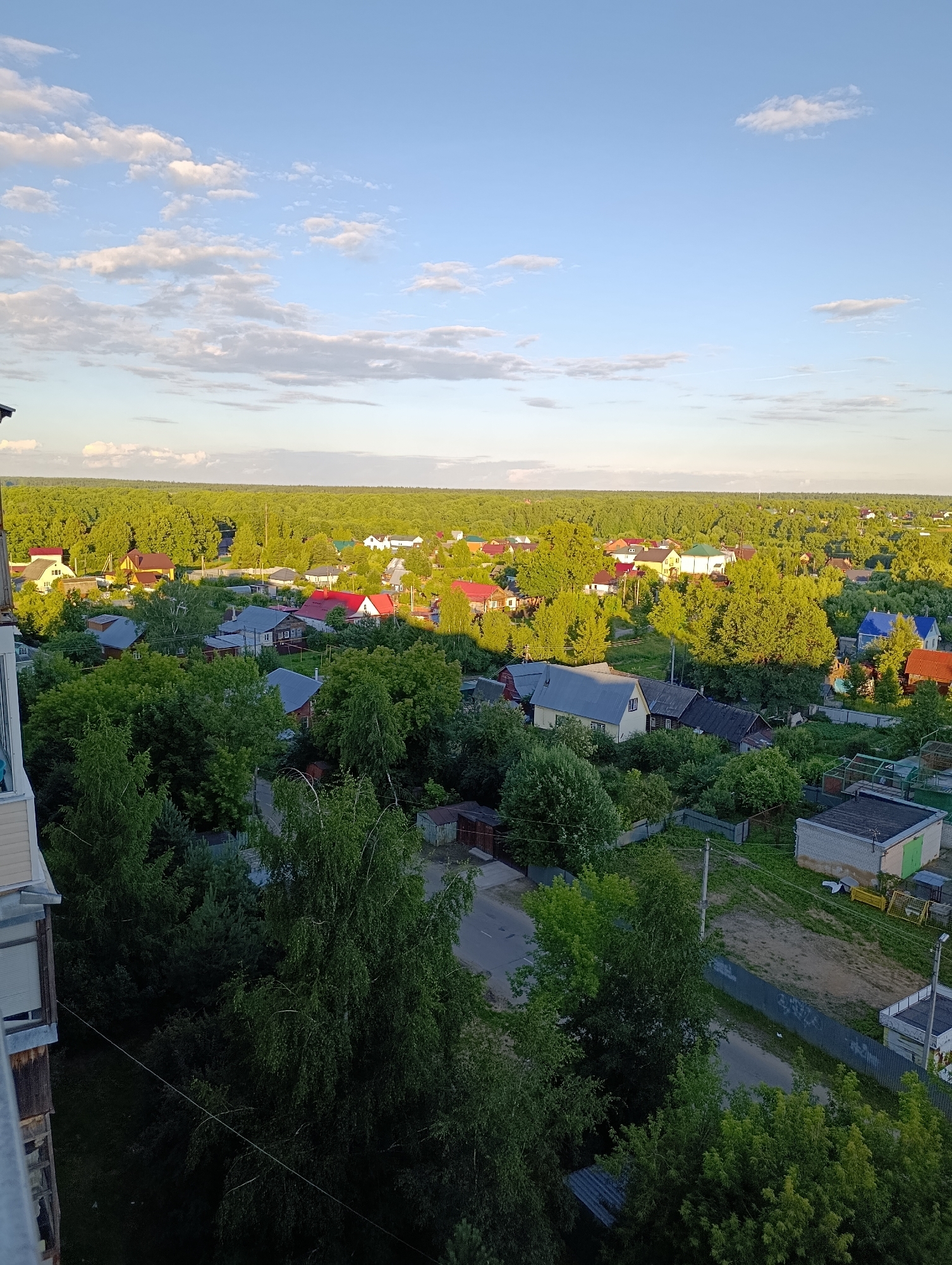 This is an aerial, eye-level, full shot of a rural village or town in Alexandrov, Russia, bathed in the warm light of a late afternoon or early evening sun. The sky above is a clear, pale blue, dotted with scattered, wispy white clouds, suggesting fair weather.
The foreground is dominated by the upper portion of a multi-story building on the left, constructed with beige siding and showing elements of windows and railings, indicating a residential structure. Below this building, lush green trees, predominantly deciduous with some evergreens, crowd the scene, their leaves catching the golden light. A dirt road or path winds through the vegetation, disappearing into the middle ground.
The middle ground reveals a cluster of houses, varying in size and architectural style. Several have bright red roofs, while others are topped with grey or darker colored roofing materials. The houses are interspersed with more trees and greenery, creating a picturesque, somewhat dense settlement. Small fences delineate property lines in some areas.
The background stretches out to a vast expanse of dense, verdant forest that appears to surround the settlement. This forest extends to the horizon, creating a natural boundary for the community. The overall impression is one of a peaceful, somewhat isolated rural living environment, characteristic of many such settlements in the Russian countryside. There are no people visible in the frame.