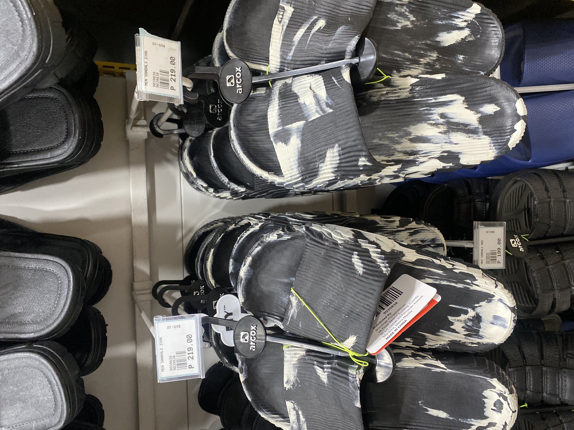 The image displays multiple pairs of sandals arranged vertically on shelves within a retail store, likely in Davao City, Philippines. The primary subjects are numerous pairs of black and white patterned slides, each featuring an abstract, cloud-like or marble-like design on a textured surface. These sandals are suspended from black plastic hangers that bear the "arcox" brand logo.
Beside and below these patterned slides, other types of sandals are visible, including solid black textured slides on the left and bottom right, and solid blue slides tucked further back on the right side of the frame. The scene suggests an activity of shopping or browsing for footwear in a retail environment.
Notable details include several price tags and product information cards. One tag clearly shows "MEN SANDALS 2306" priced at "P 219.00" (Philippine Pesos). Another tag visible reads "MEN SANDALS 902" with a price of "P 199.00." A larger tag attached to one of the patterned sandals specifies "Distributed By: Bricolage Philippines Inc.," including an address in "Marikina, Metro Manila," and indicates that the product is "MADE IN CHINA." Other visible text includes "9039838" (a product number repeated multiple times), "BG5-10," and "296006." The indoor lighting is typical of a commercial setting.