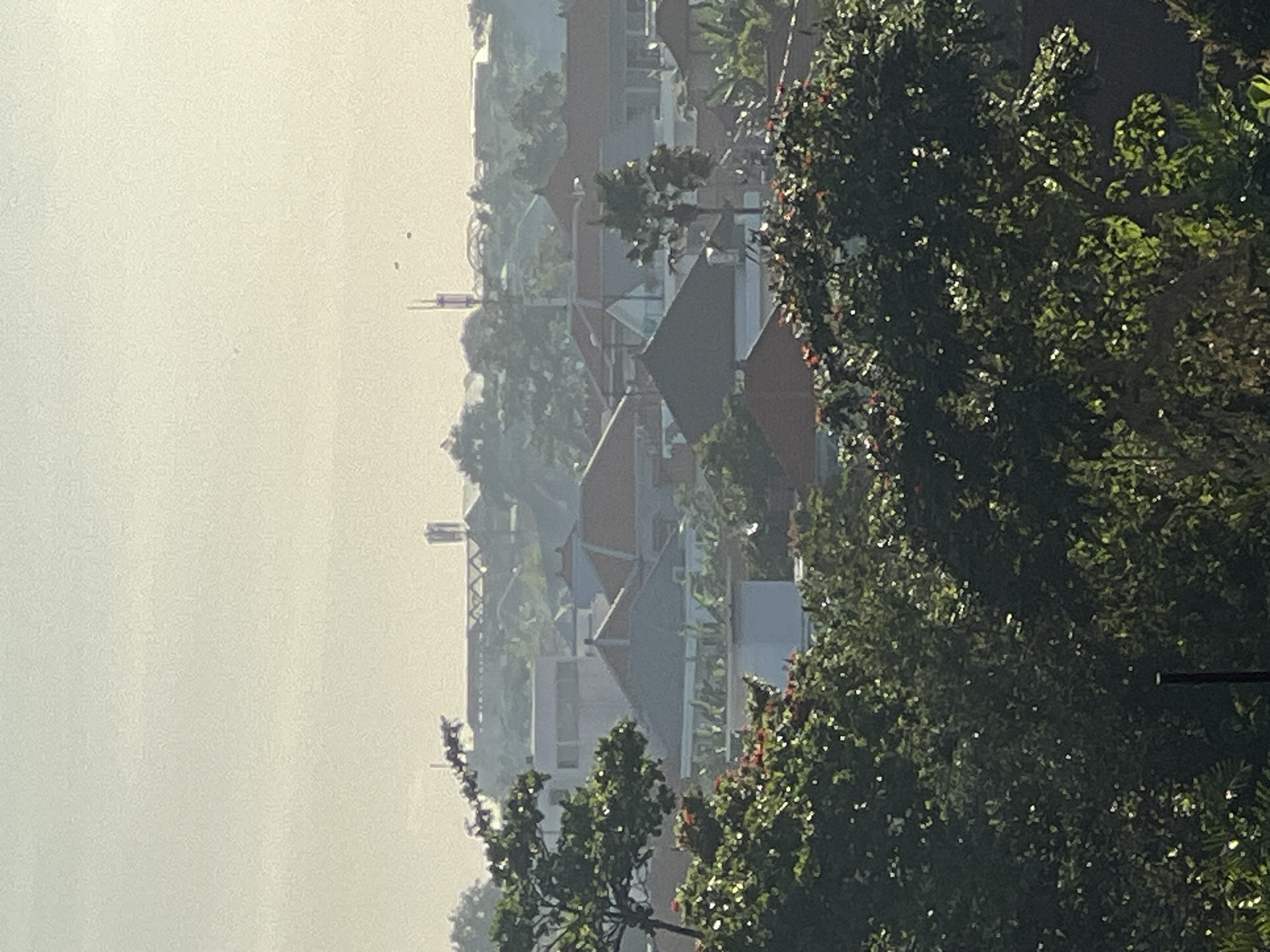 The image displays a hazy, sunlit landscape in North Kuta, Indonesia, featuring a mix of lush tropical vegetation and residential buildings. The foreground on the right is dominated by dense green foliage, likely trees, with individual leaves brightly illuminated by direct sunlight, creating a sparkling effect.
In the midground, numerous buildings with distinct reddish-brown and gray roofs are visible, scattered amongst more greenery. The architecture appears to be residential, with several structures featuring modern, geometric rooflines. Further back, taller, slender structures, possibly utility poles or communication towers, rise above the rooftops.
The background is characterized by a very light, almost monochromatic hazy sky, which obscures distant details and gives the entire scene a soft, diffused quality. Several tiny, dark specks, possibly birds, are discernible against the pale sky. The bright lighting on the foreground trees suggests it is late morning or early afternoon. No specific activities or events are taking place, and there is no visible text.