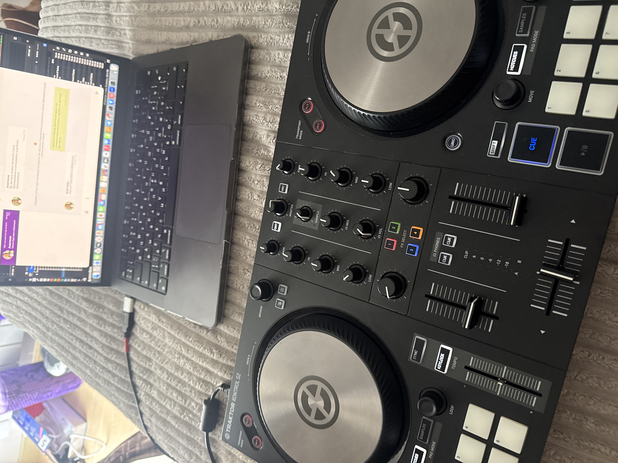 The image shows a laptop and a Traktor Kontrol S2 DJ controller on a textured surface, possibly a bed or a sofa. The laptop screen displays what appears to be a messenger app with text in a Cyrillic alphabet, suggesting a location in a region where this script is used. The Traktor Kontrol S2 controller features standard DJ controls, including jog wheels, faders, knobs, and buttons. The controller is black and silver, with various labeled functions. A cable connects the laptop and the controller. The overall environment appears to be indoors, possibly a home setting. The location is not discernible.