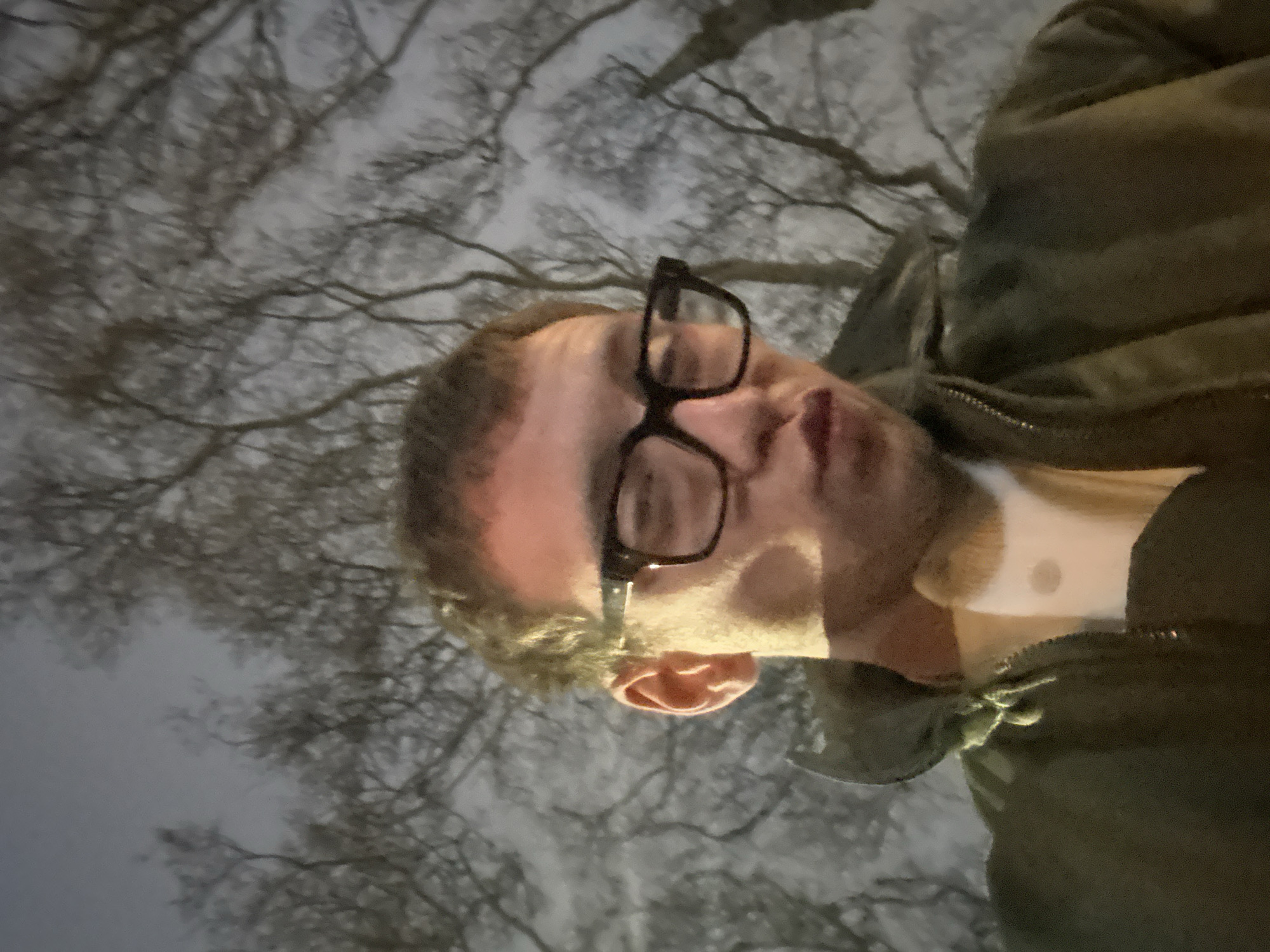 A man wearing black-rimmed glasses and a green jacket is looking at the camera. The background consists of leafless tree branches against a gray sky. The location appears to be a park or area with dense tree cover, potentially in London, United Kingdom, given the typical tree species and urban setting.