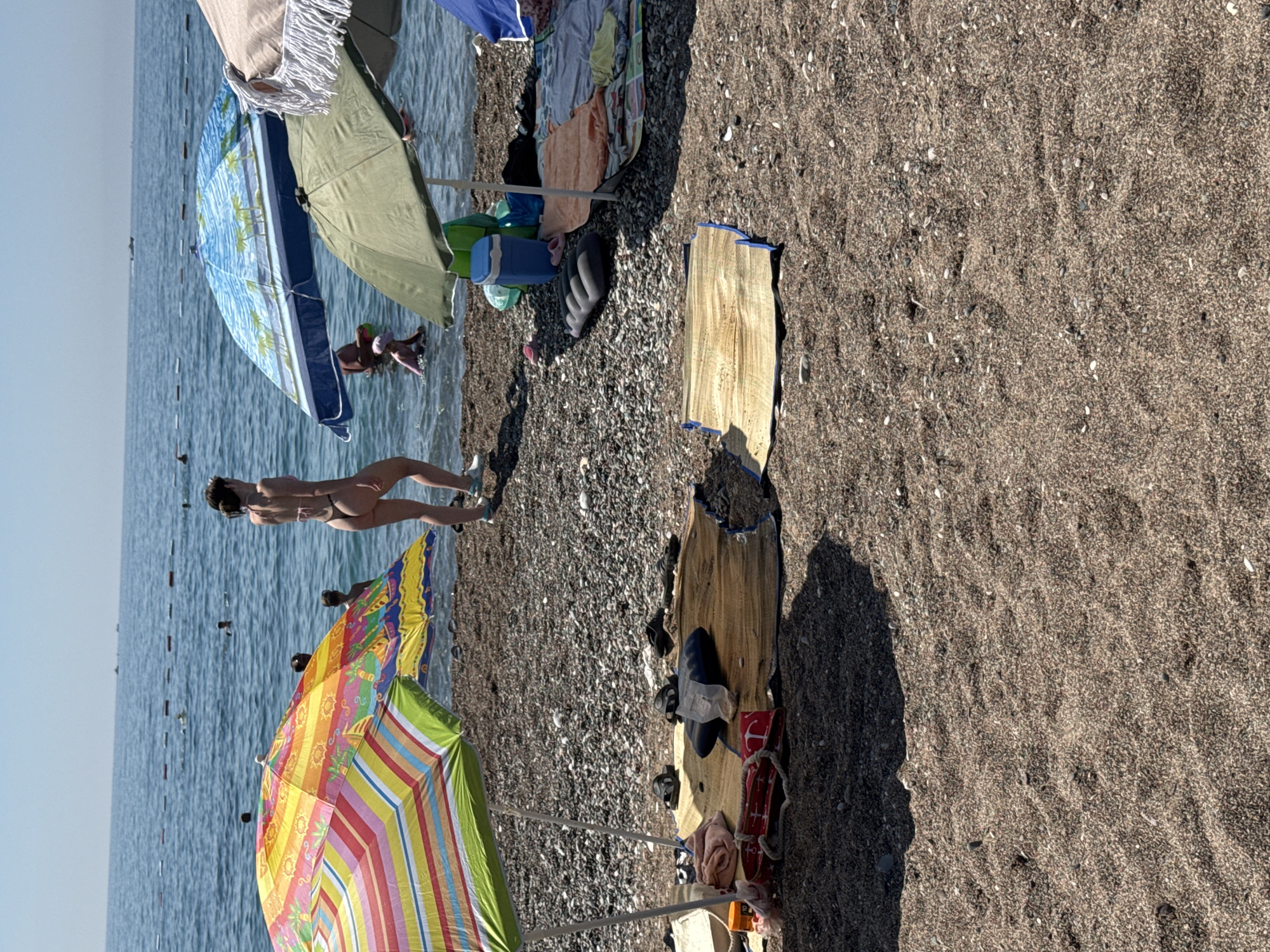 The image shows a sunny day at a pebble beach in Boreti, Montenegro. In the foreground, there are beach mats and towels laid out, with sandals and bags nearby. A colorful umbrella is set up on the left, and a green umbrella is in the center. In the middle ground, several people are visible in the shallow water. A woman in a bikini is walking along the shoreline on the right. Further out in the sea, a few more people are swimming, and a distant boat is visible. The water is calm and blue, and the sky is clear. The beach is made of small pebbles.