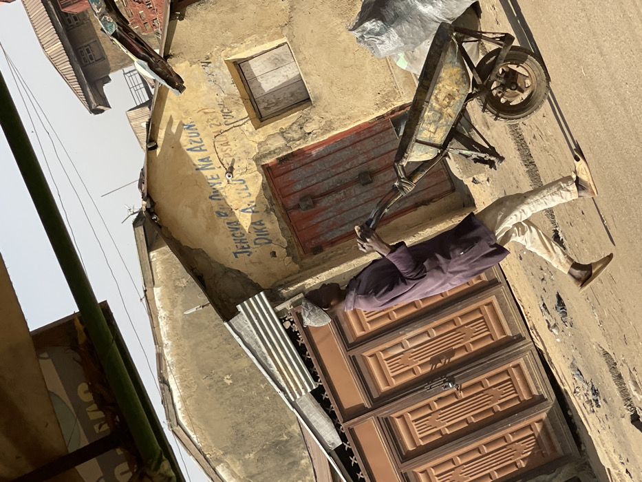 In Zaria, Nigeria, an outdoor scene depicts a male subject walking from left to right, pushing a metal wheelbarrow containing a grey bag. The man wears a light-colored cap, a dark purple long-sleeved tunic, light-colored trousers, and light brown slip-on sandals. He is positioned on an uneven, unpaved surface under direct sunlight, indicated by strong shadows.
Behind the subject, a series of weathered buildings are visible. The central building features light tan or yellowish stucco walls with visible peeling and cracks. Its facade prominently displays faded blue text: "JEHOVA BU ONYE NA AZUN", "DIKA A.ULU", and "PSALM 23:1". This building has a distressed, double-leaf, red-painted wooden door with vertical panels and a small, boarded-up window to its right. To the left of the central building, a large set of paneled brown double gates, secured with a padlock, is partially visible. Corrugated metal sheets are affixed above these gates, serving as a rudimentary awning or roofing extension. Additional building structures with corrugated metal roofs are discernible in the background. The sky is hazy light blue, and overhead, thin wires and a partial view of a green structural element are present in the upper frame. The entire image has a slight diagonal tilt.