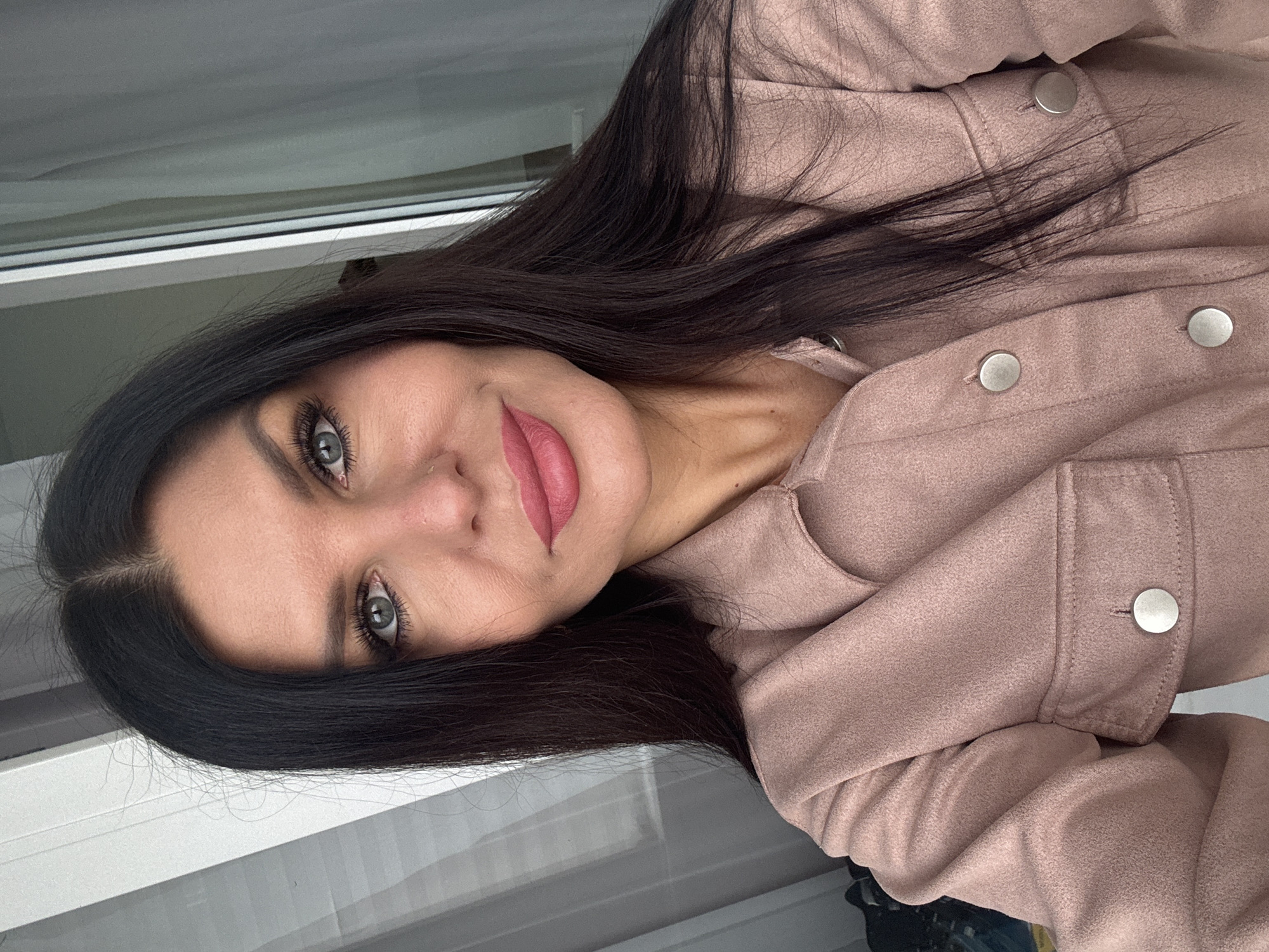 A woman with long, dark hair and light-colored eyes is seen in a close-up shot. She has fair skin and is wearing pink lipstick and a pink jacket with silver buttons. The background appears to be a window with a white frame. There is no indication of the city of Chaikovsky, Russia, visible in the image.