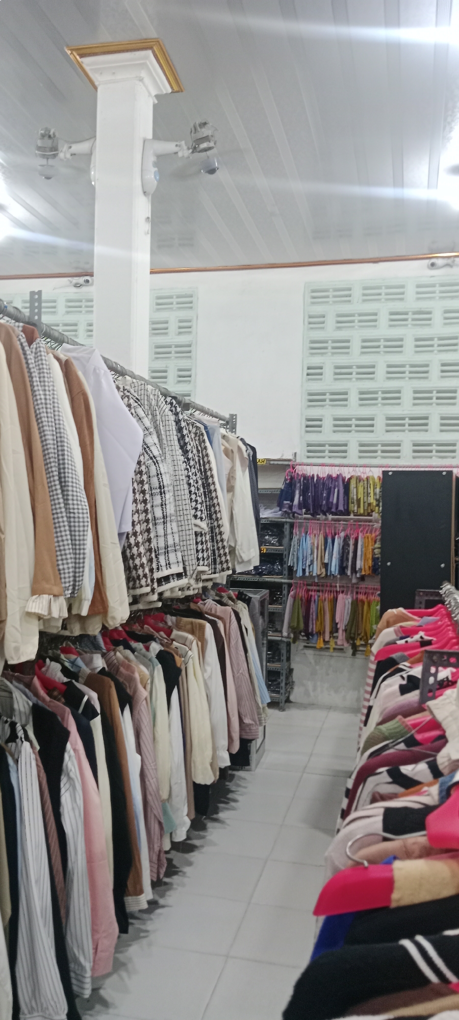The image depicts the interior of a clothing store, possibly a boutique or a section of a larger market, located in Rengel, Indonesia. The scene is well-lit, with fluorescent lighting from the ceiling casting a bright, even glow. The store features multiple racks of clothing organized by color and style.
In the foreground, to the left, a rack of women's clothing hangs. The garments are predominantly tops and jackets in various patterns and colors, including plaid, houndstooth, and solid earth tones like beige and tan. These are hung on metal hangers. Further back and to the right, additional clothing racks are visible, showcasing a wider array of apparel, including dresses and blouses, in a spectrum of colors such as pink, blue, yellow, and purple. Some of these are also displayed on hangers.
The store's architecture includes white walls and a suspended white ceiling with parallel lines. A prominent white pillar with a golden accent at its top is visible on the left side, equipped with two electric fans mounted on its sides, suggesting a need for air circulation within the store. On the walls, there are rectangular, louvered vents, a common feature in Indonesian architecture for ventilation.
The flooring is tiled in a light, neutral color, contributing to the clean and orderly appearance of the store. In the background, shelving units are visible, possibly for displaying accessories or additional stock. A black, rectangular display unit is also present on the right side of the image.
No people are clearly visible in the image, suggesting the store may be currently unoccupied or the individuals are out of frame. The overall impression is one of a well-stocked and organized retail space.