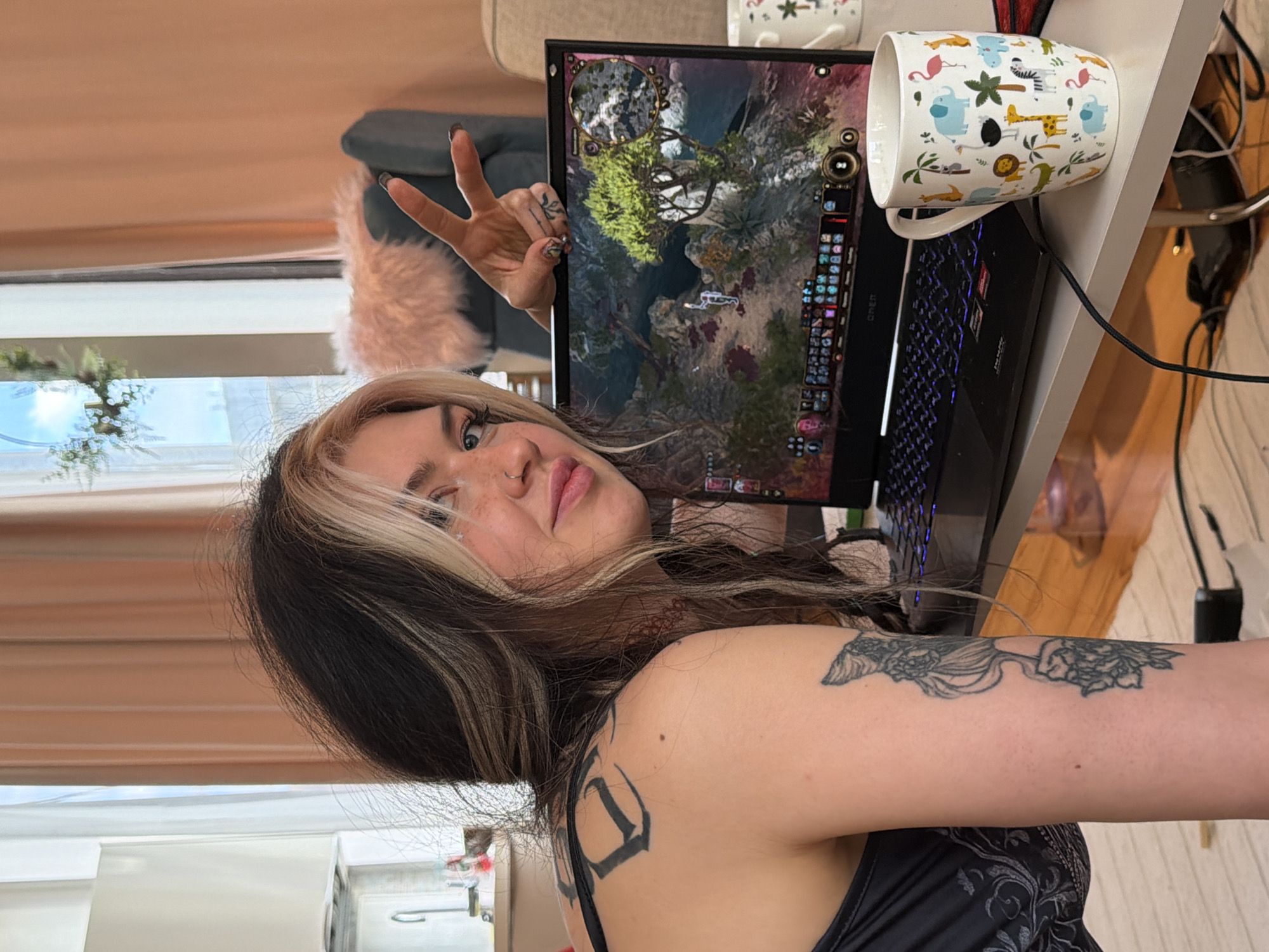 A woman with black and blonde highlighted hair is posing next to a laptop with a game visible on the screen. She is making a peace sign with her left hand and has visible tattoos on her right arm, shoulder, and fingers. The laptop is sitting on a desk next to a mug decorated with animal illustrations. A window with a view of greenery and a curtain is in the background. No specific location in Budva, Montenegro, is discernable from the image.