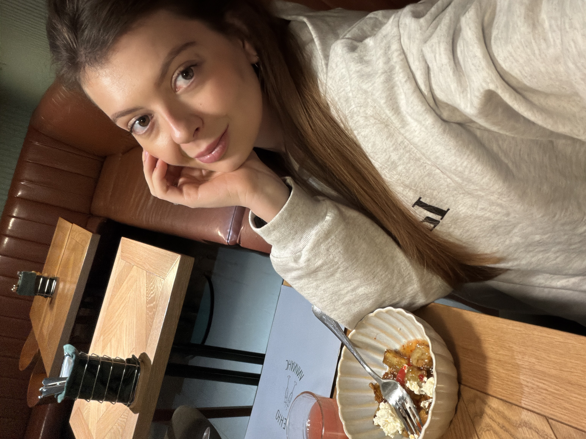 A young woman, likely in her late teens or early twenties, poses for a selfie in an indoor setting, presumably a cafe or restaurant in Khimki, Russia. She has long brown hair and is looking directly at the camera with a gentle smile, her left hand resting under her chin. She is wearing a light grey sweatshirt, which has a dark, block letter 'I' visible on the front.
The scene takes place at a wooden table, where a white, fluted bowl containing a fruit-based dessert with whipped cream and a fork is prominently displayed. Beside it, a clear glass holds a pink beverage. A light blue menu or placemat, adorned with an illustration of two champagne glasses and mirrored Cyrillic text that appears to read "АМАНАЧ" and "ОННО," is also on the table.
The background features a brown leather booth, suggesting comfortable seating. Another wooden table with a black metal cutlery holder, containing silverware wrapped in teal napkins, is visible to the left. The artificial, somewhat dim lighting indicates it is likely evening or night. The woman's calm and pleasant expression conveys a sense of contentment during her visit.