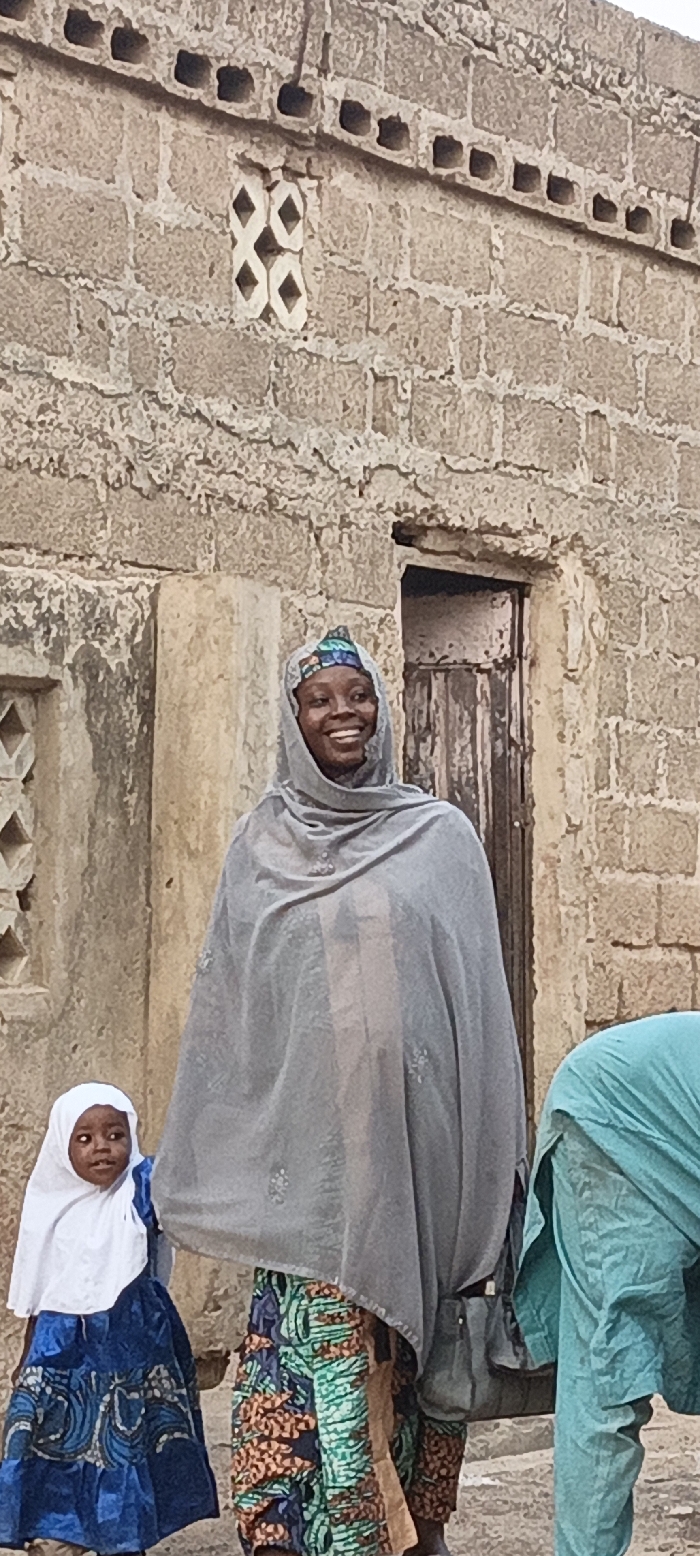 This outdoor portrait captures a vibrant street scene in Rangaza, Nigeria, featuring a smiling woman, a child, and a partially visible third person against a rustic stone building.
In the center, a woman grins broadly, her face radiating joy. She wears a grey head covering over a colorful, patterned cap, a long grey outer garment, and a brightly patterned wrapper or skirt in hues of green, blue, and orange. A dark shoulder bag hangs from her left side. To her left, a young child, likely a girl, stands wearing a white head covering and a blue and white patterned dress. The child gazes to her left with a neutral, inquisitive expression. On the far right, another person is partially visible, dressed in a light blue or turquoise garment, appearing to be bent over or obscured.
The background is dominated by a sturdy, light-colored stone wall constructed from rough-hewn blocks. Decorative elements are embedded in the wall, including a white geometric pattern resembling connected diamond shapes and a horizontal band of small square openings. A dark, aged wooden door is visible behind the main woman. The bright, even illumination suggests it is daytime. The scene conveys a sense of community and daily life, with the main subject's happy expression being a notable detail. No visible text is present.
