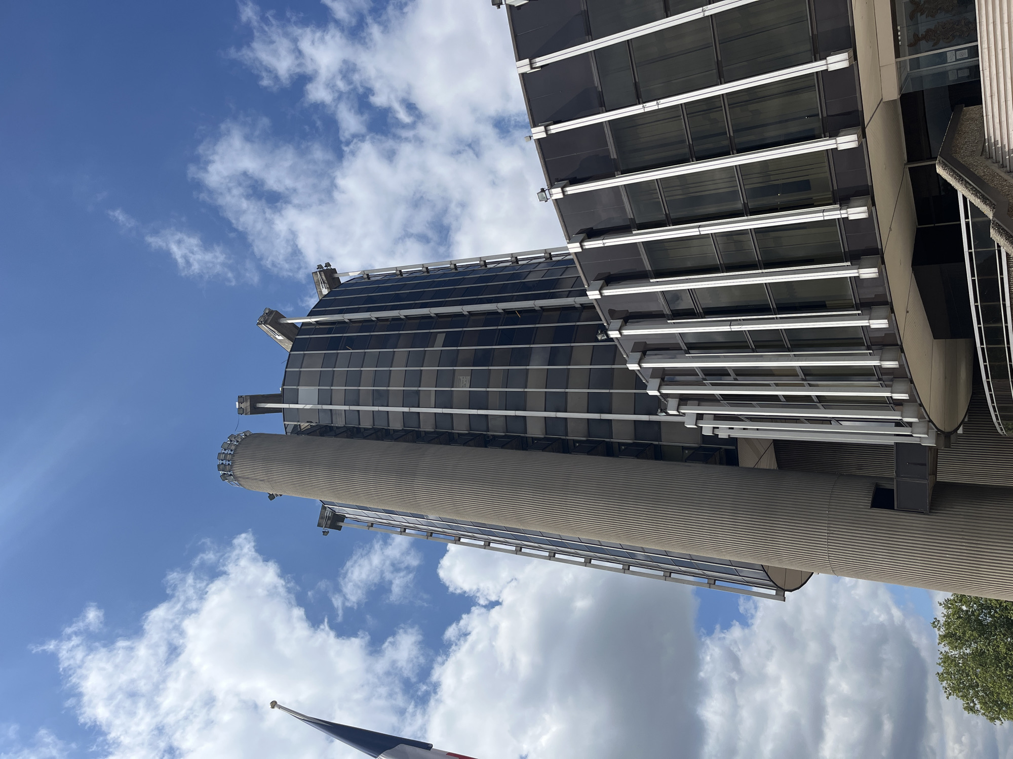 The image shows a low-angle shot of the Palais de Justice in Créteil, France, under a bright, cloudy sky. The building features a prominent, cylindrical concrete tower with vertical ribbed detailing. Adjacent to the tower is a glass-and-steel structure with a dark, reflective facade. The building's architectural style appears to be modernist. A French flag on a flagpole is visible on the left side of the frame. The environment suggests an outdoor urban setting on a daytime. There are no people visible in the image.