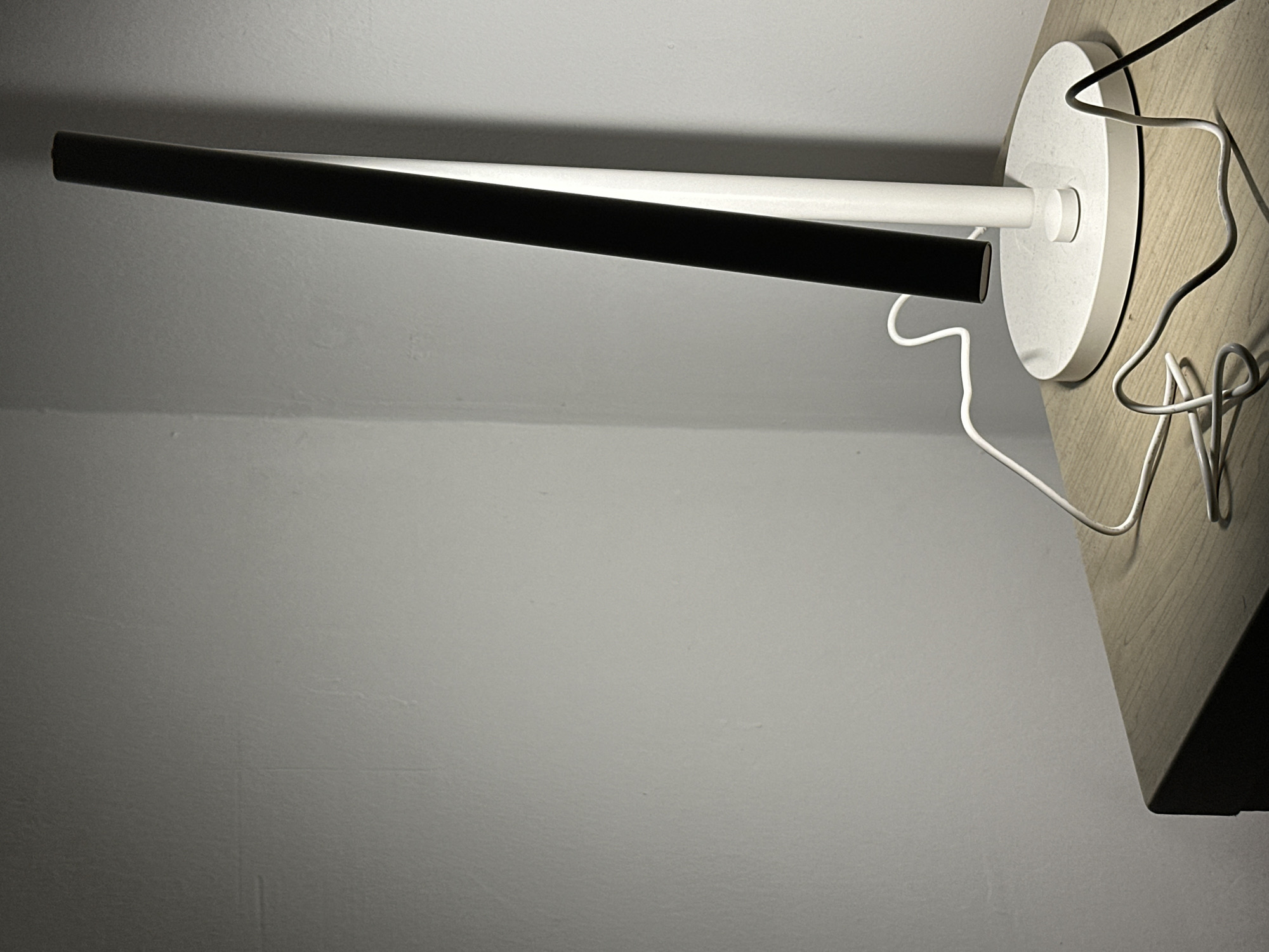 In a room located in Ramat Gan, Israel, a modern, minimalist desk lamp stands on a light-colored wooden surface, likely a table or nightstand. The lamp features a flat, circular white base, a vertical white cylindrical support, and a black rectangular light source angled upwards from the support, emitting light. A white electrical cable emerges from the lamp's base, coiling loosely on the wooden surface, and a partially visible black cable is also present to the right of the lamp. The background consists of two light gray walls meeting at an interior corner, with the left wall appearing dimmer than the right due to lighting. The scene is illuminated primarily by the lamp, casting shadows on the walls and surface. No individuals are present in the frame.