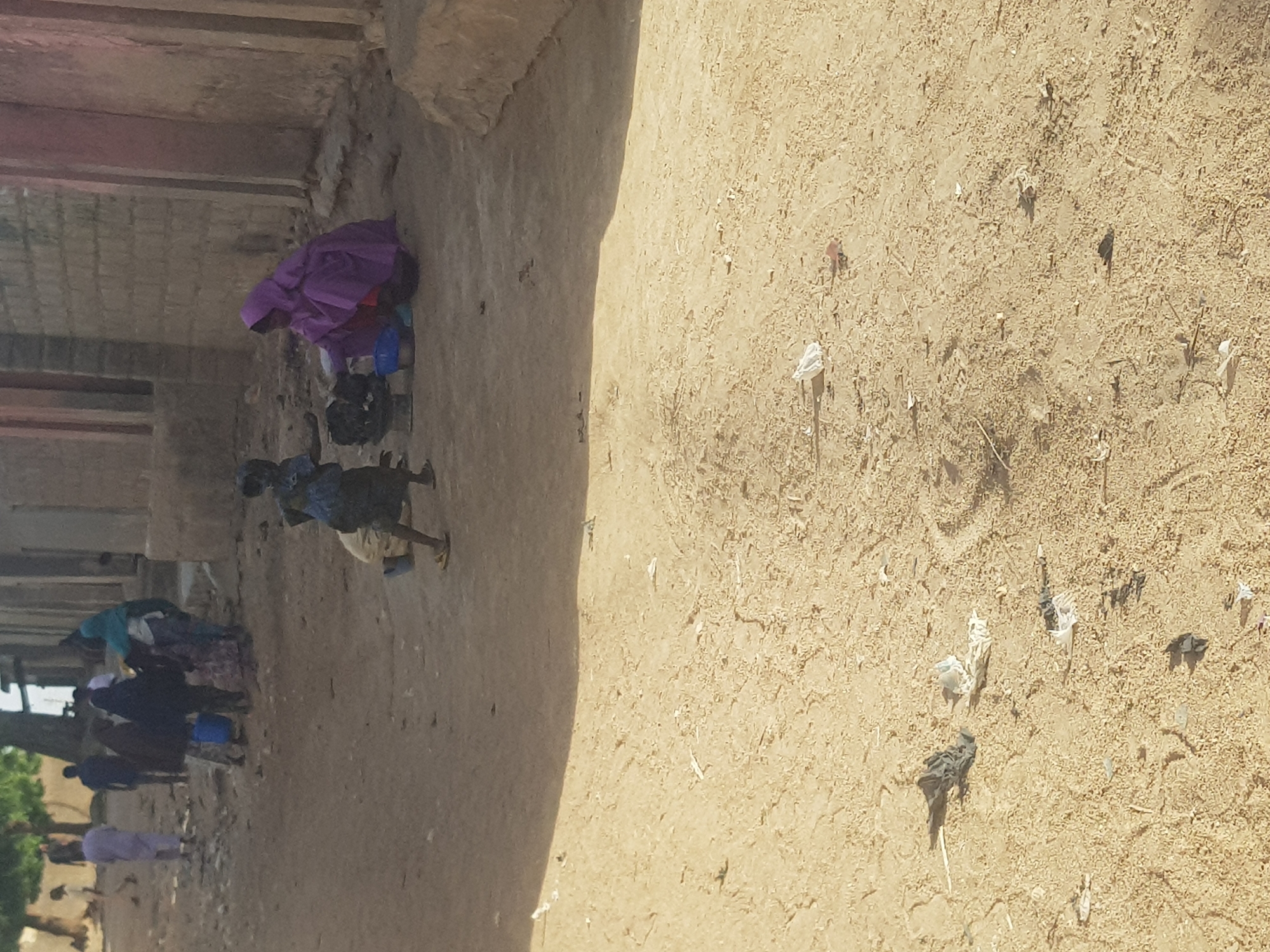 The image depicts a dirt road in what appears to be a residential area, likely in Dabawa, Nigeria. Several individuals, possibly women, are gathered along the side of the road. Some appear to be carrying items on their heads or working on the ground. The ground is covered in sand and debris. The background shows buildings with weathered walls. The lighting suggests it is daytime.