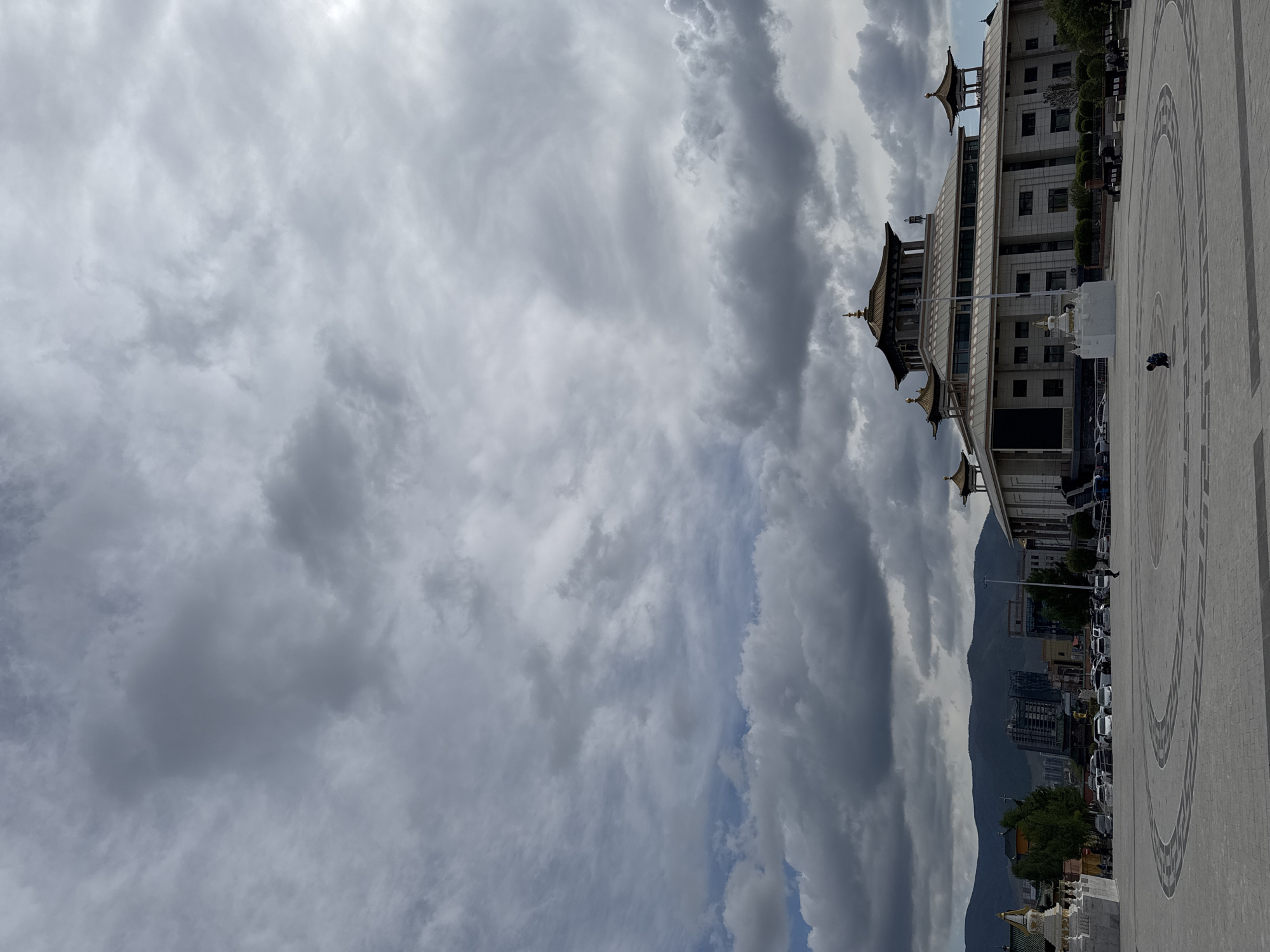 A building with a multi-tiered roof stands adjacent to a gray paved plaza, with outlines of circular designs, located in Ulaanbaatar, Mongolia. Mountains form a backdrop. Clouds dominate the sky. Cars are parked between the building and the mountain. A person is visible in the plaza.