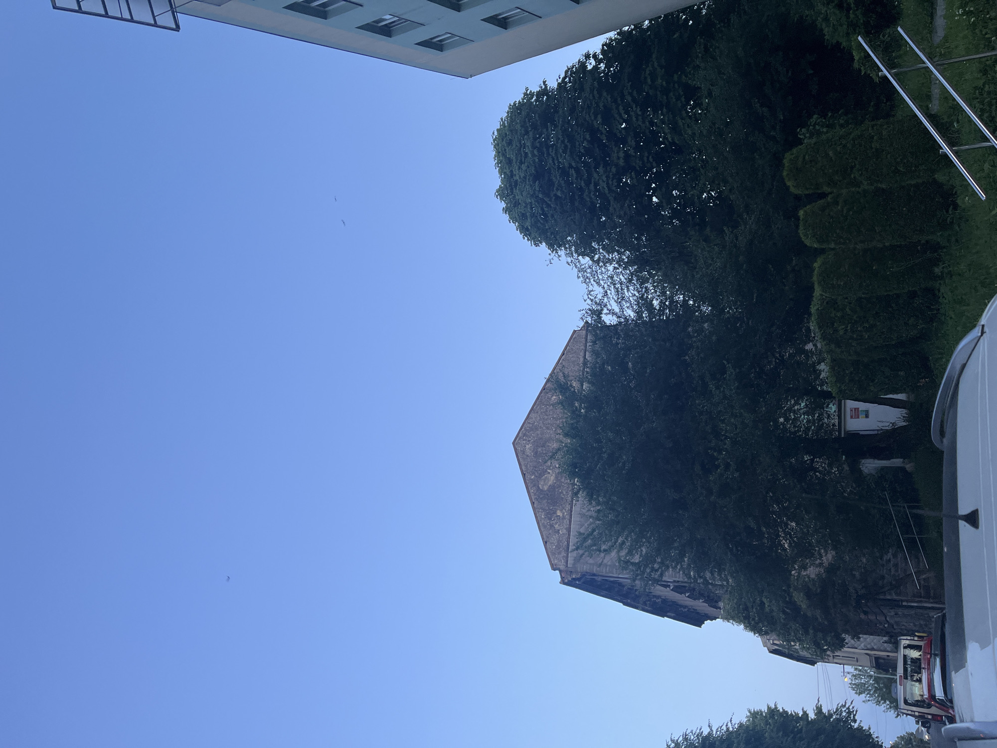 The image shows a low-angle view of a building and trees against a clear blue sky. The building appears to be made of stone with a weathered texture and a steep, angular roof. Lush green trees surround the building, with some casting shadows on its facade. To the left, a portion of a modern building with multiple windows is visible, suggesting an urban or suburban setting. There are also some metallic structures in the background, possibly part of an antenna or scaffolding, and a white vehicle partially visible on the right side. The lighting suggests it is daytime, with the sun casting a soft glow. No people are visible in the frame, and the overall impression is one of peaceful coexistence between nature and architecture.