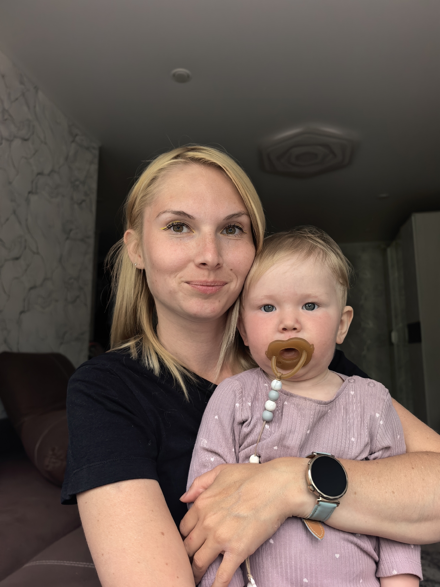 A blonde woman in a black t-shirt holds a baby wearing a pink onesie with a pacifier. The woman has yellow eyeliner and a watch on her left wrist. The baby has blue eyes and chubby cheeks. They are in an indoor setting with a patterned wall and a dark ceiling. The background suggests a living room or apartment, with a glimpse of a couch. The image is a portrait-style photo, focusing on the two subjects. The specific location within Sterlitamak, Russia, cannot be determined from the visual information provided.
