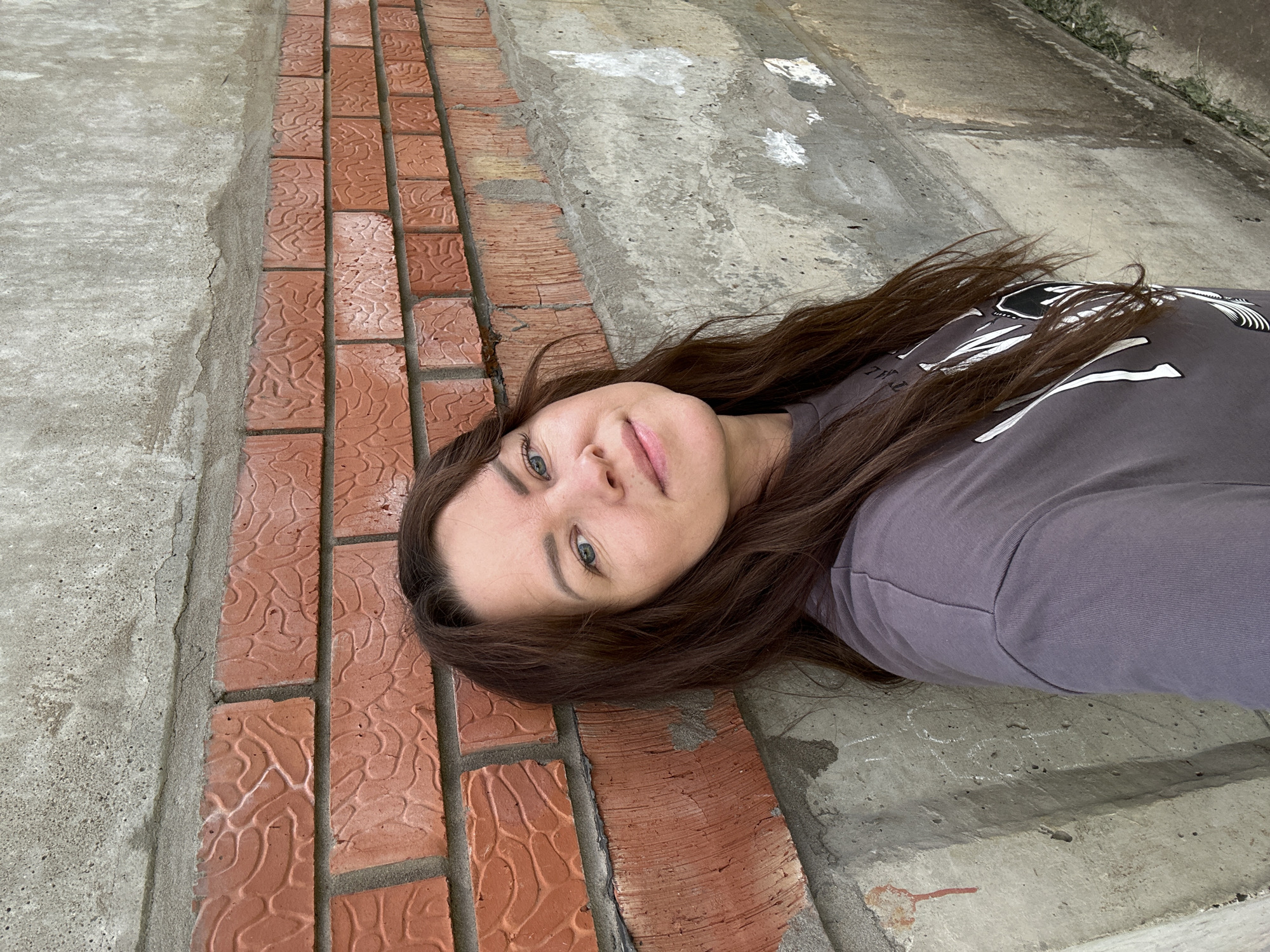 The image shows a woman leaning against a brick and concrete wall outdoors. The woman has long, dark brown hair and is wearing a dark grey t-shirt with a white graphic design. She is looking towards the camera with a slight smile. The wall is made of red brick with a textured pattern, and the concrete section below it is rough and shows signs of wear and some white markings. The background suggests an outdoor urban environment. The location is identified as the city of Chaikovsky, Russia.