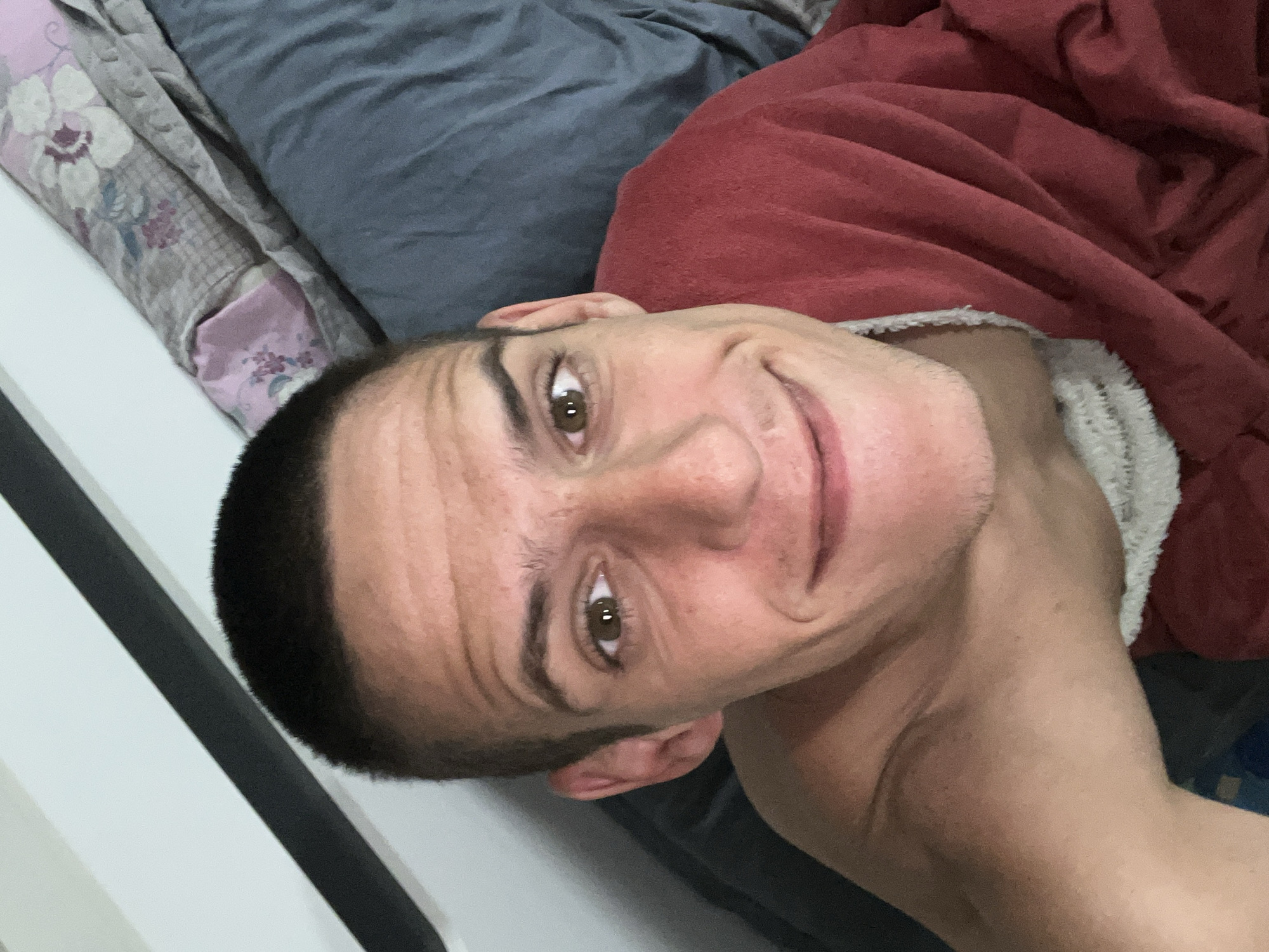 A man with short, dark hair and light skin is lying in bed, covered partially by a red blanket. He has brown eyes and is smiling slightly. The bedding includes a patterned pillow and a blue sheet. The environment suggests a bedroom setting. The location of the photo is not discernible from the image and therefore cannot be confirmed as Sofia, Bulgaria.