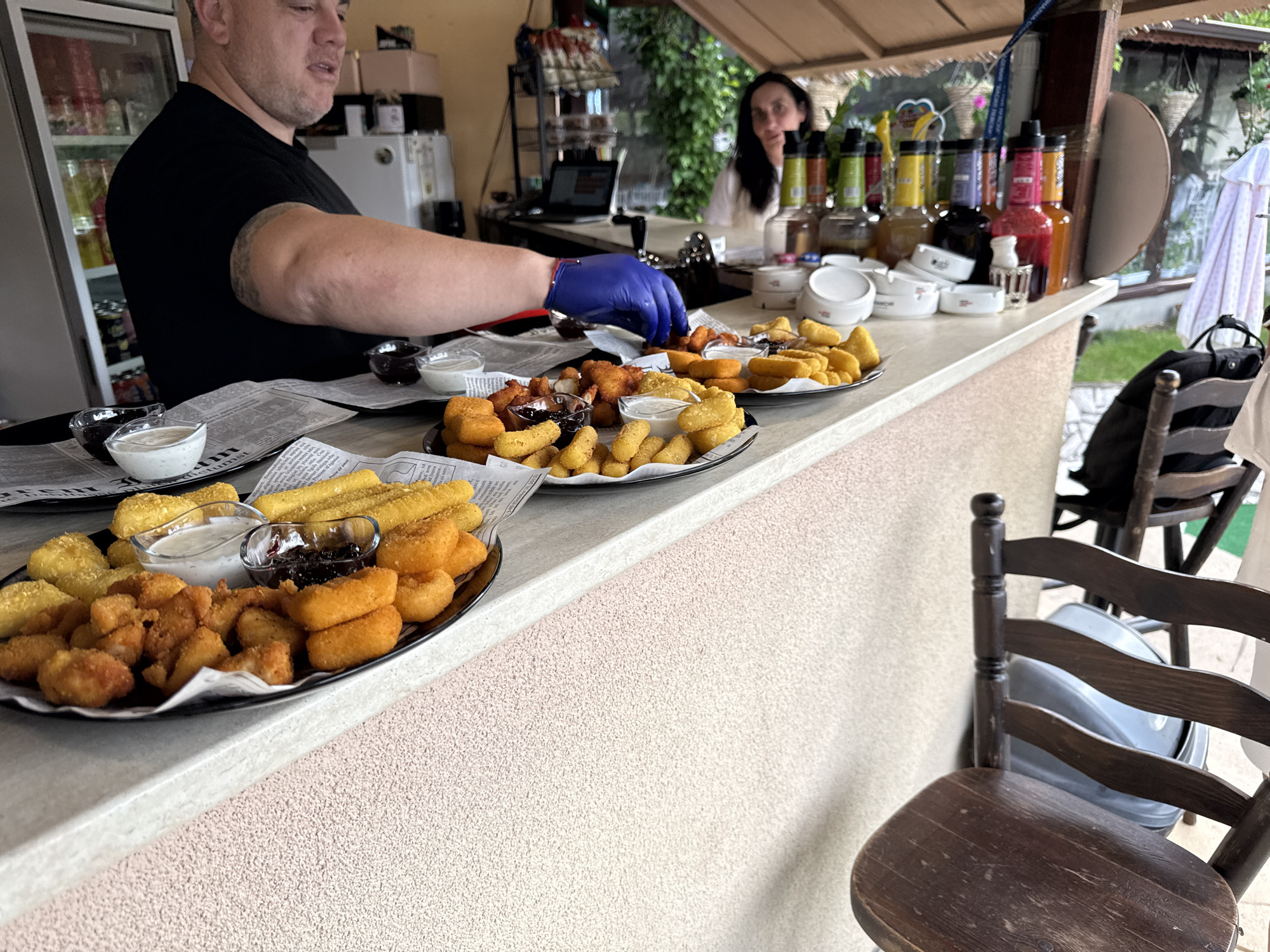 This image captures an outdoor food preparation and serving area, identified as being in Radlovtsi, Bulgaria. The primary focus is a male individual in a black short-sleeved shirt, positioned on the left, with his right arm extended over a counter. He wears a blue disposable glove on his right hand and appears to be arranging or preparing food on one of several platters. Visible on his left bicep is a partial tattoo. His gaze is directed downwards towards the food.
In the mid-ground, a woman with dark hair is partially visible behind the counter, looking towards the right. She is wearing a light-colored top. The counter surface in the foreground is light grey, transitioning to a light pink textured surface directly beneath it.
Spread across the counter are multiple dark, circular platters, each lined with what appears to be newspaper print. These platters hold various fried food items, including golden-brown nuggets, sticks, and rings. Each platter is accompanied by small glass bowls containing two types of dips: a white creamy sauce and a dark, possibly jam-like or savory sauce. At least two such platters are prominently displayed.
Behind the counter, a cluttered area includes a white refrigerator visible on the far left, partially obscured shelves with various items, and numerous bottles of colorful liquids, likely syrups or mixers, lined up on a shelf behind the man and woman. Several white ashtrays are also present on the counter in this area. A laptop is visible on a surface further back.
The environment suggests an outdoor or semi-outdoor setting, with lush green foliage and a thatched or rustic wooden overhang in the background, indicative of a patio or garden bar. A dark wooden chair with a curved back is positioned in the lower right foreground, facing away from the counter. The lighting is bright and even, consistent with daylight.