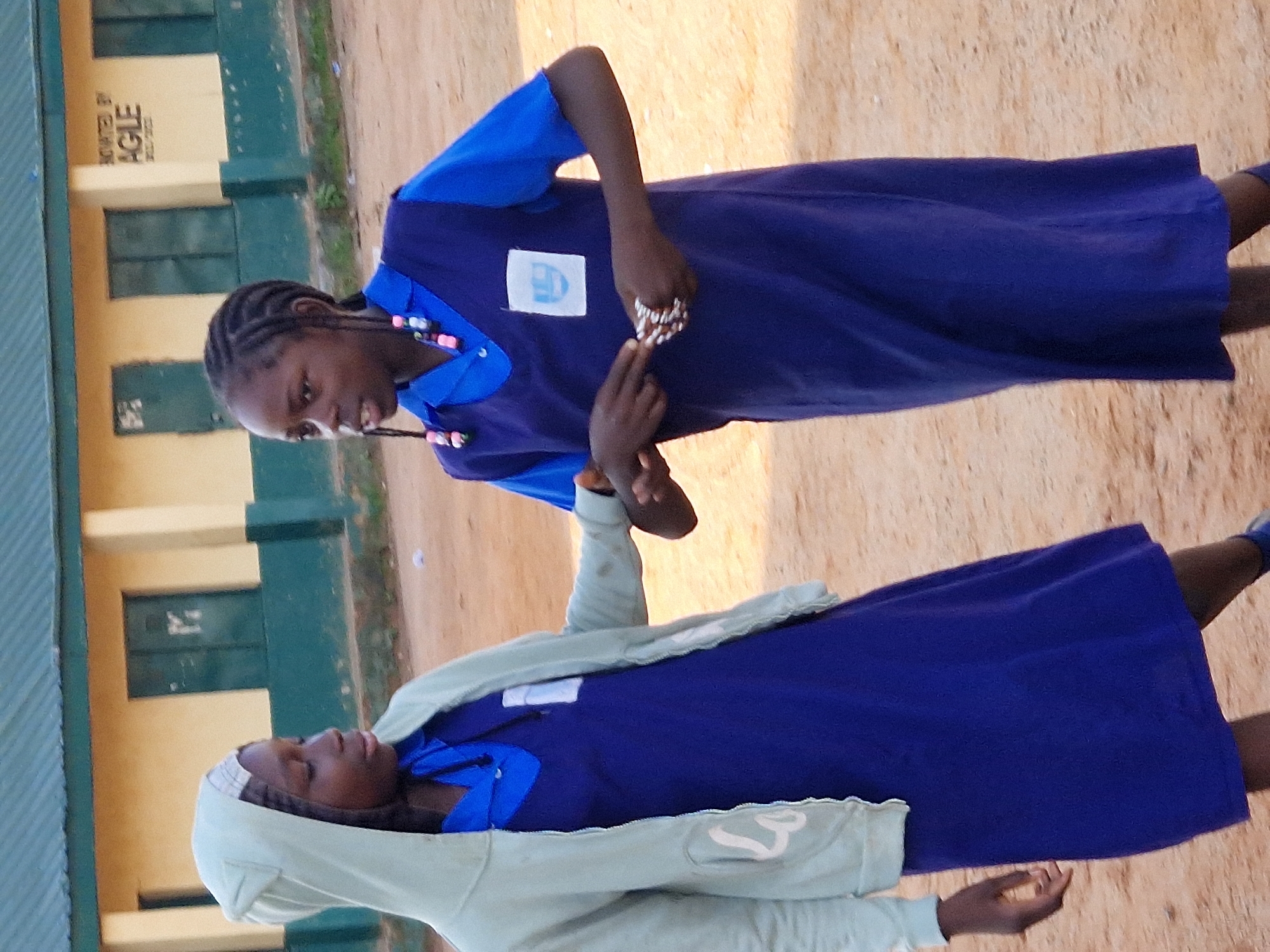 Two young Black girls, likely students, stand outdoors in front of a light yellow building with a dark green roof. The girl on the left wears a light blue hooded jacket over a dark blue dress and a headscarf. The girl on the right wears a dark blue dress. She has braided hair adorned with pink and blue beads. She is holding a bracelet made of small, light-colored beads in her hands. The ground is sandy and brown. The building has dark green doors and windows. Text is visible on the building which reads "OWED BY AGILE." The scene is likely within the city of Kabong, Nigeria, as indicated by the presence of a school building and its architecture.