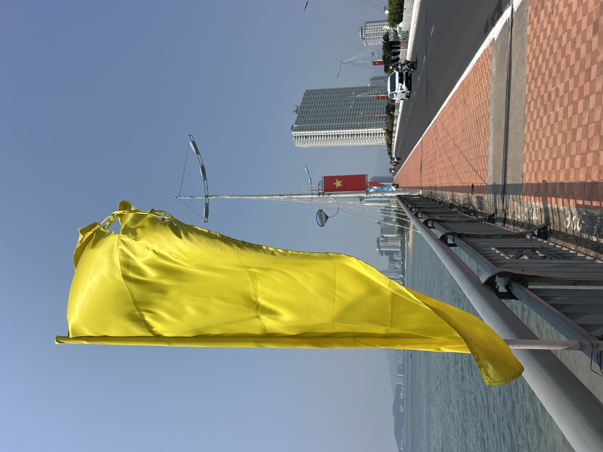 The image shows a yellow flag waving in the wind on a bridge railing. The bridge spans a body of water, and in the background are buildings and a city skyline, identifiable as Bắc Nha Trang, Vietnam. A Vietnamese flag is visible further down the bridge, along with other decorative elements such as tall flagpoles with reflective circular discs and arching supports. A brick-paved sidewalk borders a road with vehicular traffic, including a white car. The sky is a clear blue. The image appears to be taken during daylight hours.
