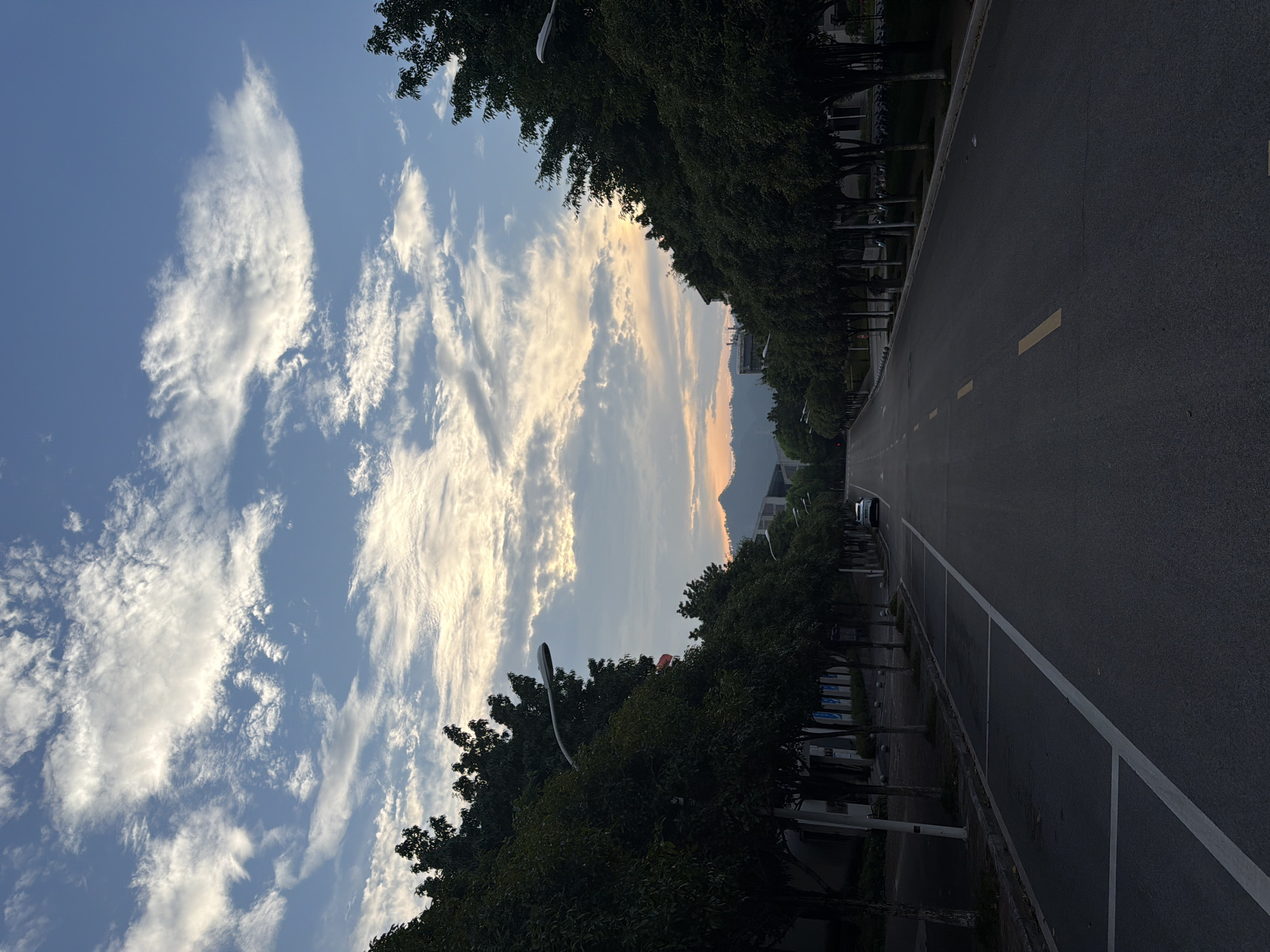 The image depicts a road lined with trees in Jiangjin, China, during what appears to be either sunrise or sunset. A straight, paved road stretches into the distance, marked with yellow and white lane dividers. On either side of the road, a dense row of mature trees with dark green foliage creates an avenue effect. In the background, mountains are visible against a sky filled with scattered white and grey clouds illuminated by warm, golden light, indicating twilight. A single white car is parked on the right side of the road. Streetlights are positioned along the sidewalk on both sides. Buildings are partially visible behind the trees on the left. The overall impression is a serene, orderly urban landscape.