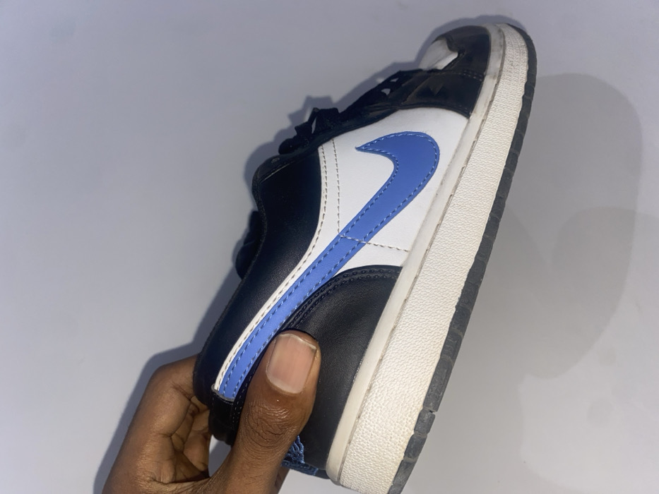 A hand holds a black, white, and light blue Nike Air Jordan 1 Low sneaker. The shoe has a black toe box, white side panels, and a light blue swoosh. The midsole is white with a black outsole. The shoe appears used, with slight creasing and wear on the sole. The background is a light grey or white.