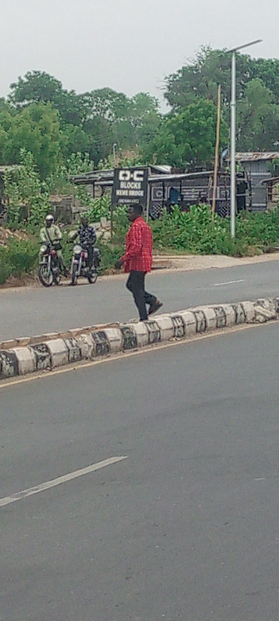 In Adankolo, Nigeria, a man in a red patterned shirt and dark pants walks along a concrete median separating the road. Two men sit on motorcycles on the roadside, partially obscured by vegetation. Behind them is a sign that reads "OC BLOCKS MEME BRIDGE." A metal structure is in the background and there are trees and a lampost.