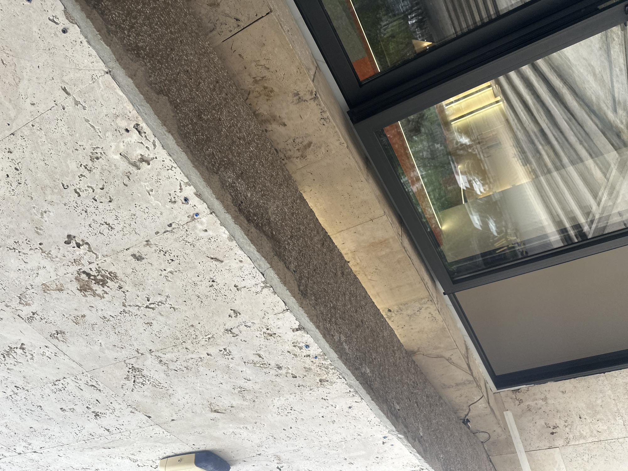 A low-angle, close-up shot depicts a portion of a building exterior, potentially in Yerevan, Armenia, although this is not explicitly confirmed visually. The architecture features light-colored, textured stone cladding and a dark-framed window. The window reflects an interior space with visible lighting fixtures and possibly draped fabrics or curtains. A linear element, possibly a beam or ledge, runs diagonally across the frame, exhibiting a rough, aggregate texture on one side and a smoother surface closer to the window. At the bottom left, a small, partially visible object suggests a mechanical or electrical component. The overall composition focuses on the textural contrasts and the interplay between the architectural elements and the reflected environment.