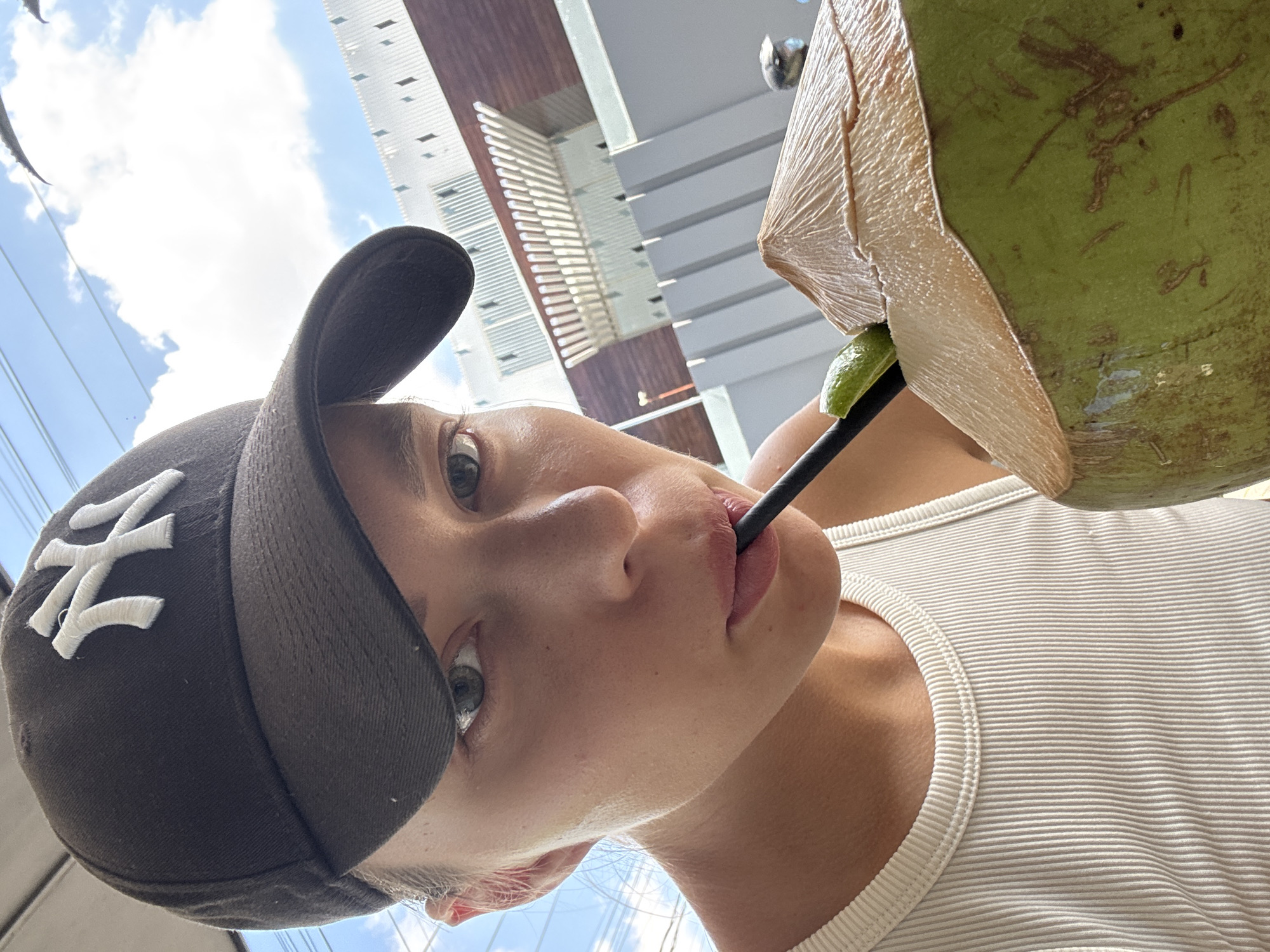 A young woman wearing a baseball cap with the New York Yankees logo and a white ribbed tank top is drinking from a fresh coconut with a black straw and a wedge of lime. She is standing outside with a partially cloudy sky and tall buildings in the background. The scene is likely in an urban area, possibly in North Kuta, Indonesia.