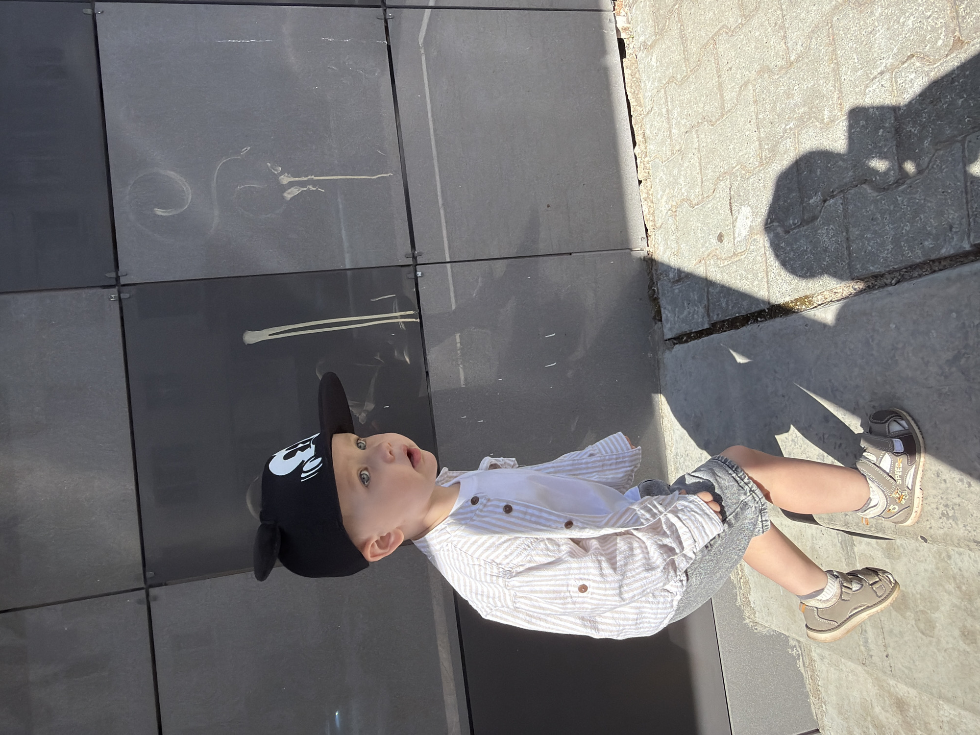 This is a portrait-oriented, eye-level, full-body shot of a young child, likely a toddler, seated outdoors on a paved surface. The child is wearing a black baseball cap with Mickey Mouse ears and a white t-shirt with a striped button-down shirt over it. The child is also wearing denim shorts and closed-toe sandals. The child is looking upwards and to the left with a slightly open mouth, appearing to be engaged with something or someone off-camera. Their expression suggests curiosity or mild surprise.
The background consists of two distinct textures: a dark, matte, tiled surface on the left and a lighter, textured, grey stone or concrete paving on the right. Sunlight casts a distinct shadow of the child and their surroundings onto the paved area, indicating it is daytime with clear weather. The light suggests it is likely late morning or early afternoon. There are some faint white markings on the dark tiles, possibly water stains or graffiti. The child's attire and the sunny environment suggest a casual outdoor activity, perhaps a moment of play or rest during a walk. There are no visible signs or text in the image. The overall impression is a bright, everyday moment captured outdoors.