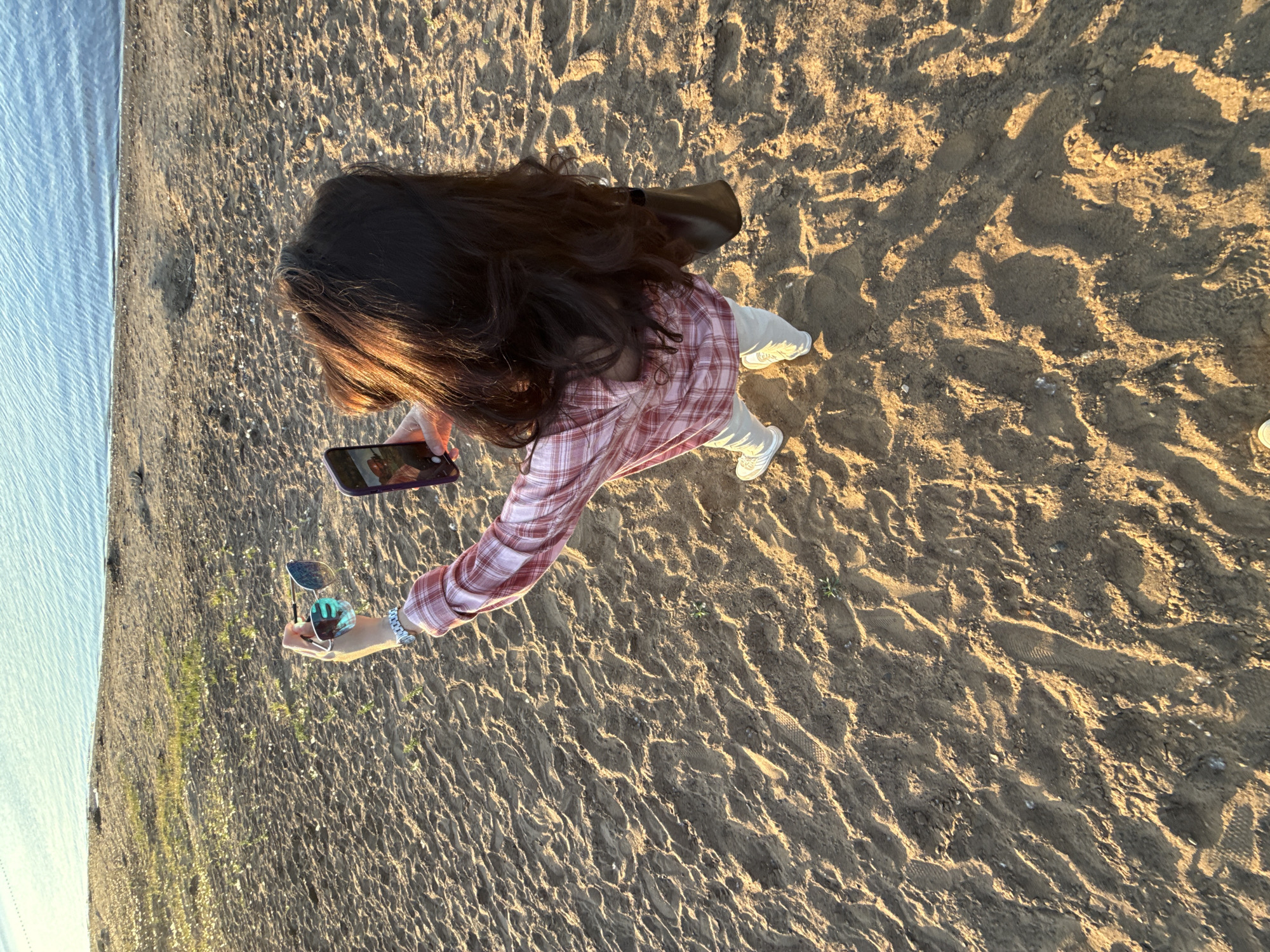 The image depicts a scene on a sandy shore in the city of Chaikovsky, Russia. The foreground and midground consist of light-brown sand, marked with numerous footprints and exhibiting distinct ripples and shadows cast by low-angle sunlight. Sparse green vegetation protrudes in isolated patches from the sand. A large body of calm, blue water, likely a lake or river, occupies the background.
One person, with long, dark brown hair illuminated by the sun, is positioned in the center-right of the frame, viewed from a high-angle perspective. The individual wears a red and white plaid long-sleeved shirt, light-colored pants, and white athletic shoes. A silver-toned watch is visible on their left wrist, and a dark-colored shoulder bag hangs from their right shoulder. The person is holding a dark-cased smartphone in their right hand, angled upwards as if taking a photograph or selfie, with a reflection visible on the screen. In their left hand, they hold a pair of blue-mirrored sunglasses. The scene is bathed in warm, golden light, indicative of either sunrise or sunset.