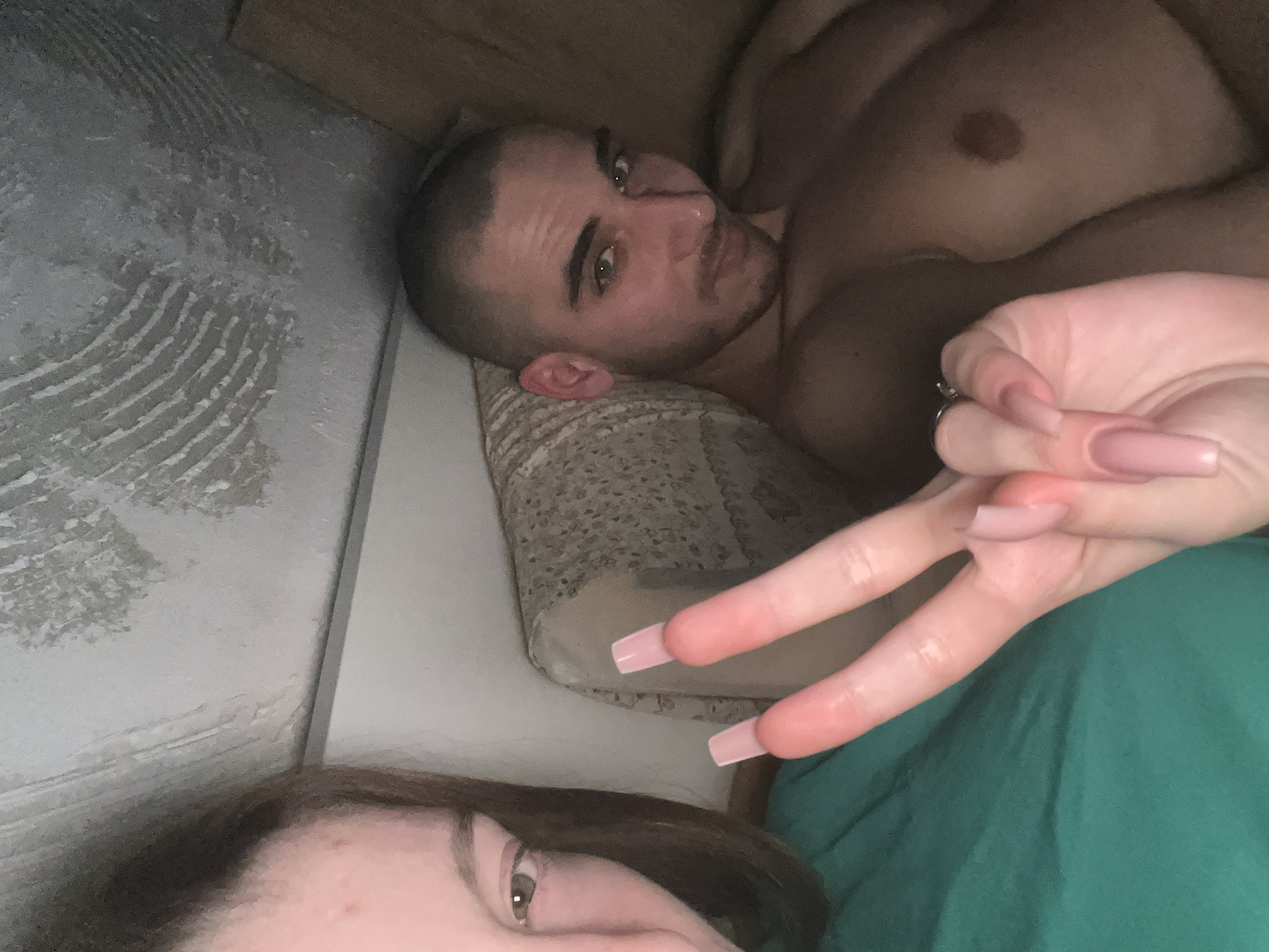 A selfie captures a shirtless man with a shaved head lying on a patterned pillow and gazing at the camera. Beside him, a woman partially visible in the lower frame, displaying a peace sign with her hand, adorned with long, pink acrylic nails. They are lying on a green surface, possibly a bed or blanket. The location is unknown; there is no evidence to suggest this image was taken in Varna, Bulgaria.
