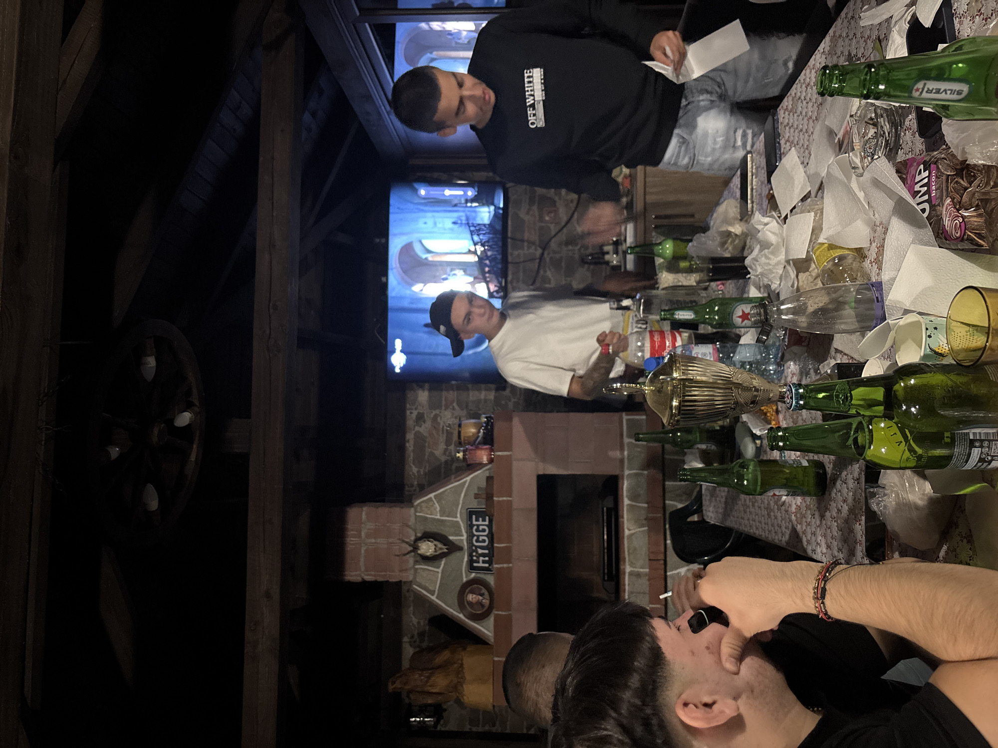 In Zlatograd, Bulgaria, a dimly lit, rustic outdoor covered patio area appears set for a casual gathering. Four young men are present. In the foreground, a man wearing a black t-shirt and a bracelet on his left wrist is seen from the side, looking towards the right. Beside him, another man in a black t-shirt is visible from the back, with a distinctive haircut. Across the table, two other men are standing. One, wearing a white t-shirt and a black baseball cap, is looking towards the left, holding a bottle. The other, dressed in a black long-sleeved shirt with "OFF WHITE" printed on it and blue jeans, is gesturing with his right hand while holding a piece of white paper in his left.
The central focus is a table laden with items. Several green glass bottles, a plastic bottle of Coca-Cola, and various glasses are scattered across a patterned tablecloth. Used paper napkins are also present, along with an ashtray containing cigarette butts. A large trophy cup stands prominently on the table. Behind the table, a brick fireplace is visible, adorned with a deer skull and a sign reading "HYGGE." A television screen displaying what appears to be an interior scene with arches and statues is mounted above the fireplace. The ceiling is constructed of exposed wooden beams, contributing to the rustic ambiance. The overall scene suggests a social event with drinks and light refreshments.